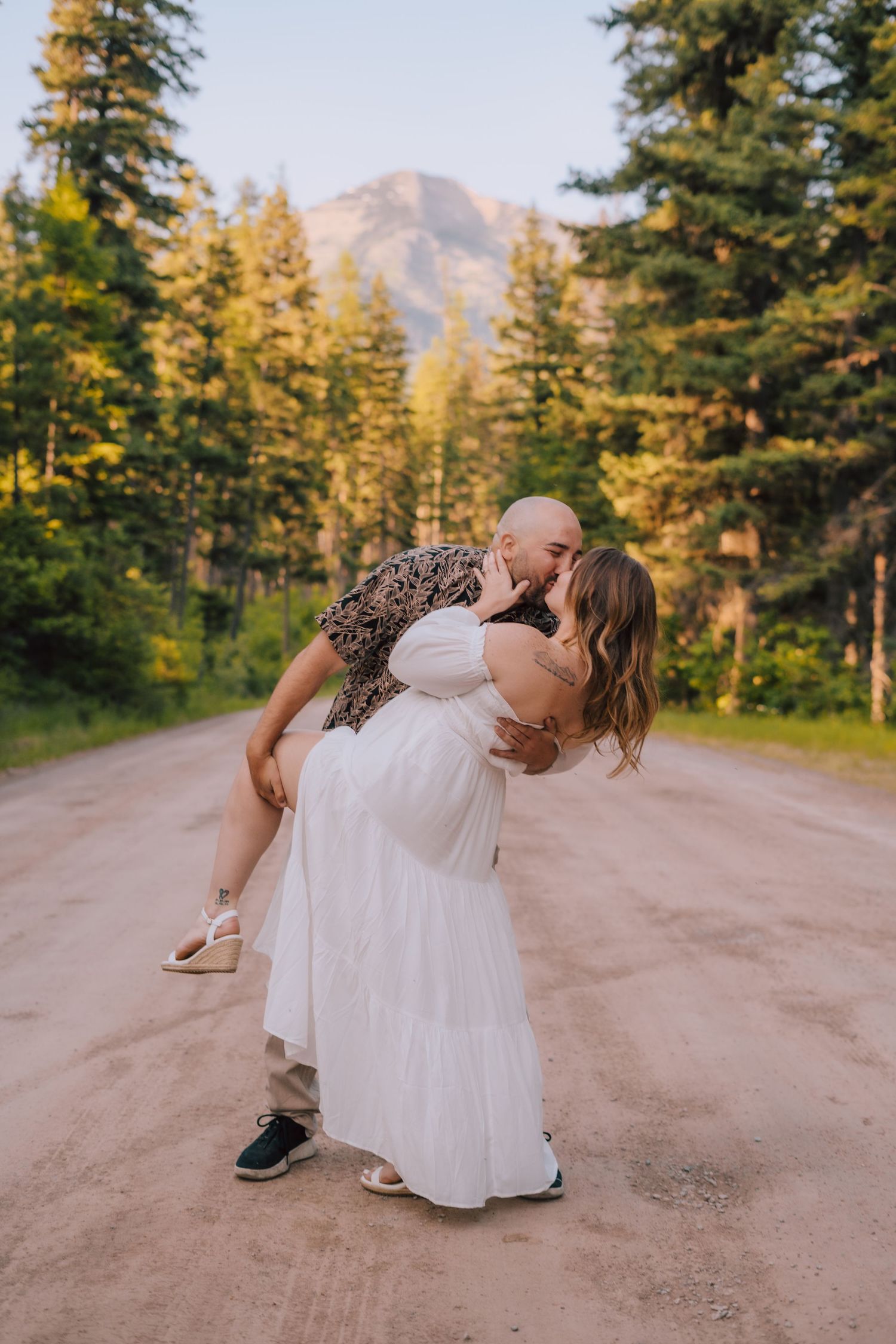 A romantic kiss on a dirt road surrounded by evergreen trees with a mountain peak in the distance at sunset.