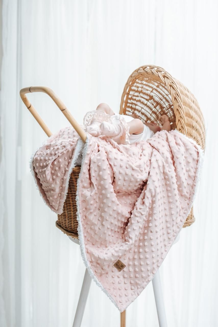 Pink minky blanket and baby booties displayed in wicker bassinet with bamboo handle against white curtain background.