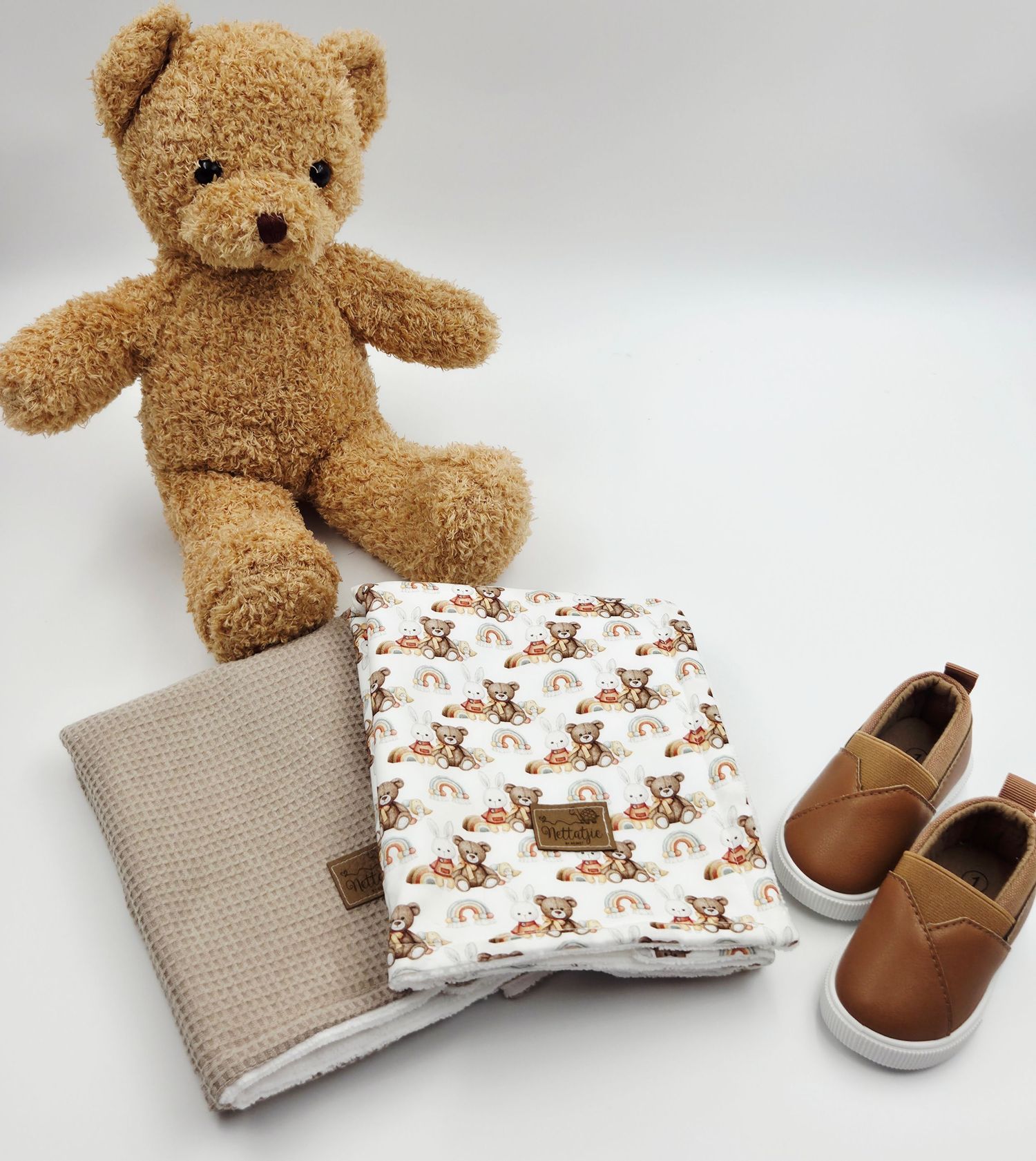 Brown teddy bear with baby blankets and small brown leather shoes arranged on white background.