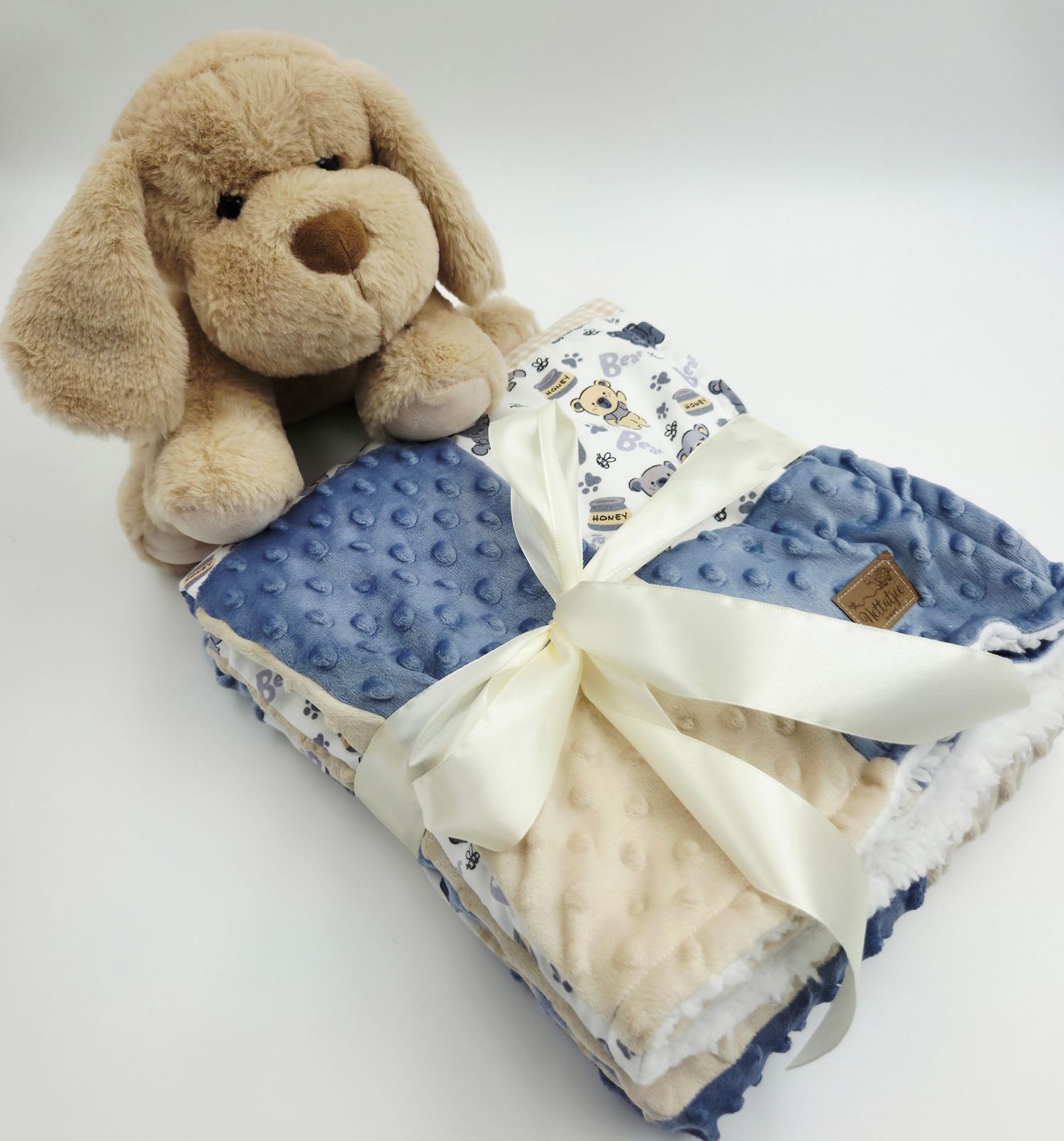 Plush puppy toy resting on folded blue and beige patchwork blanket with white ribbon.