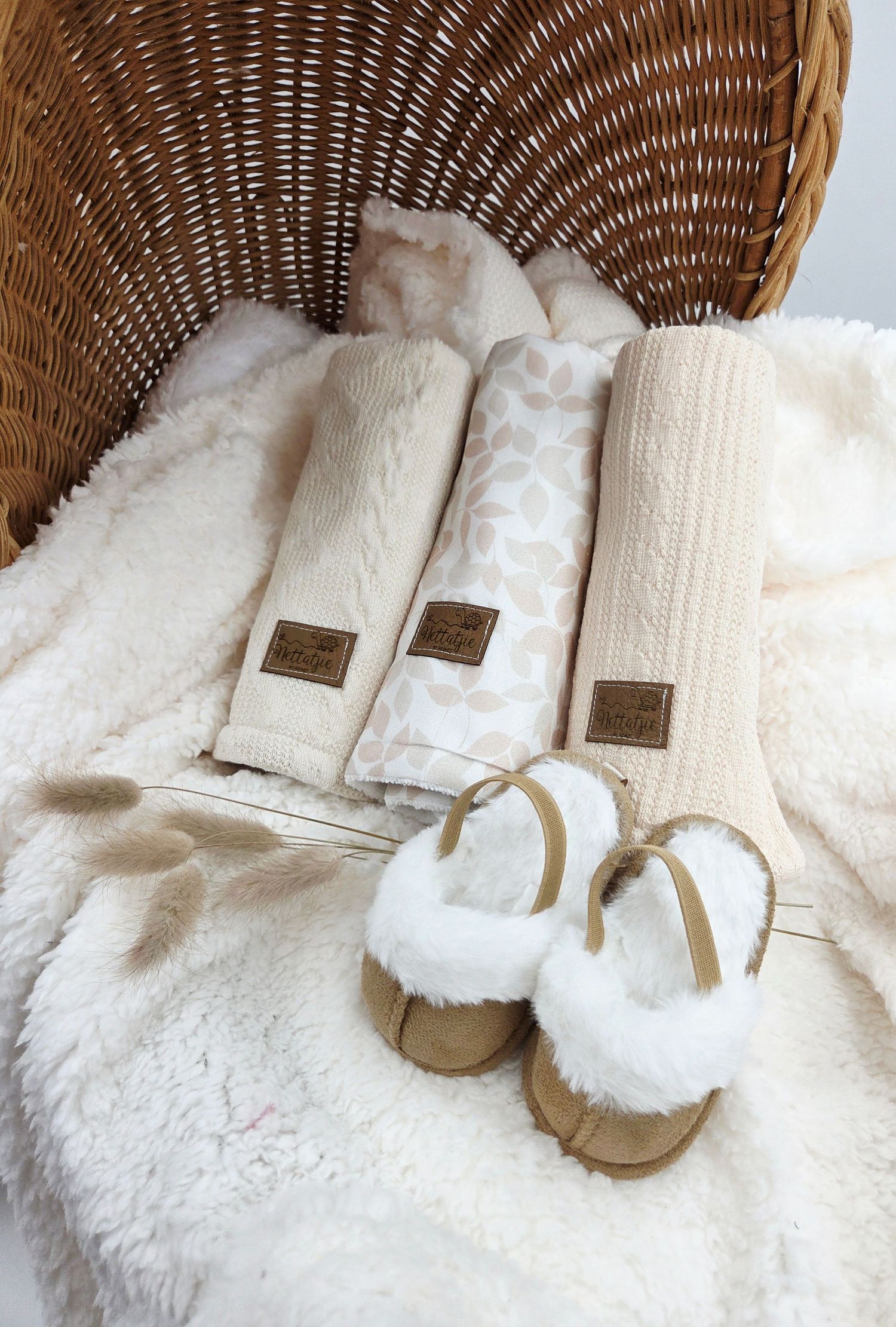Neutral baby shoes and blankets arranged in wicker basket with plush white fabric.