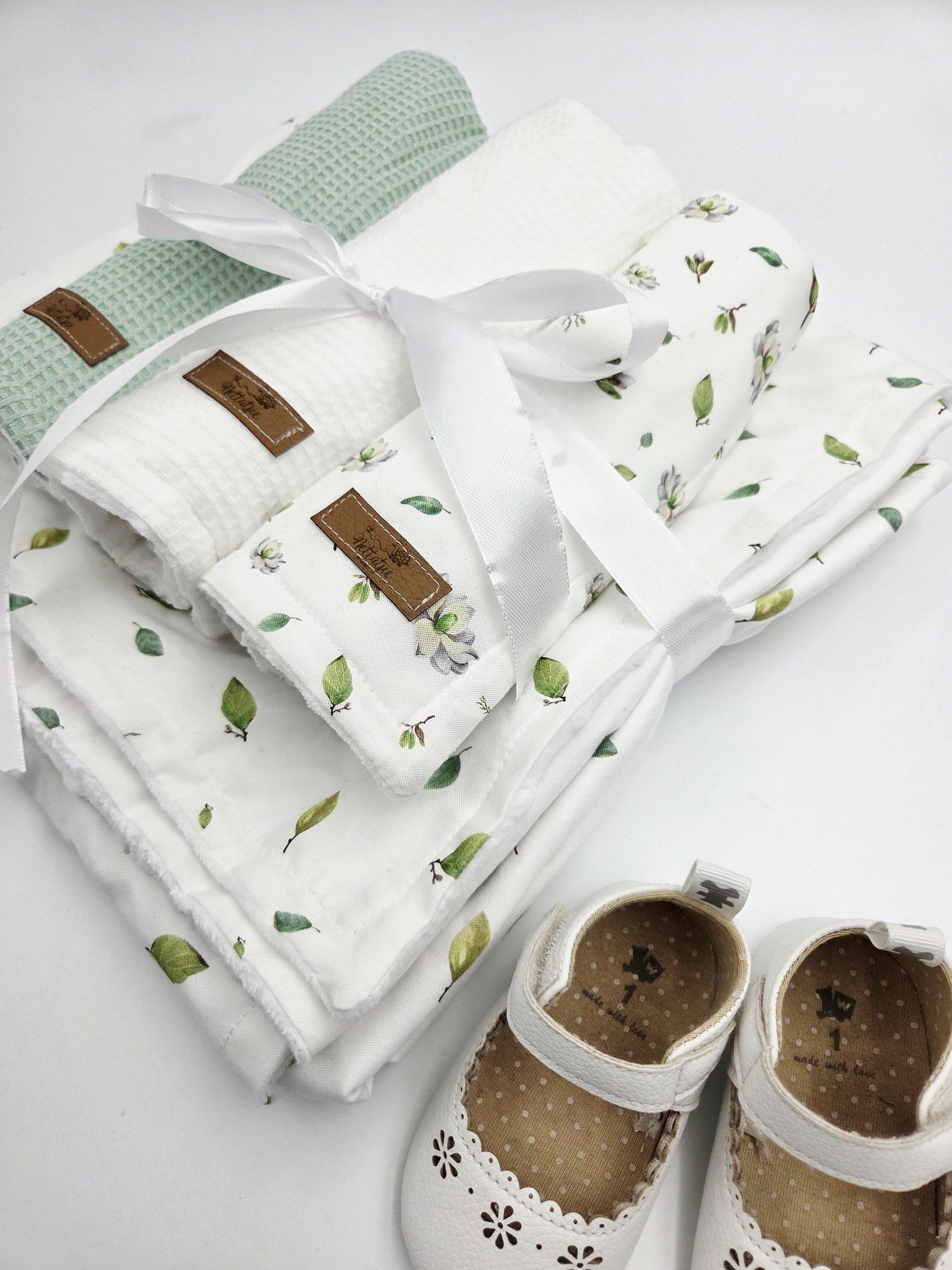 Mint green and white textured baby blankets with botanical print arranged with white baby shoes.