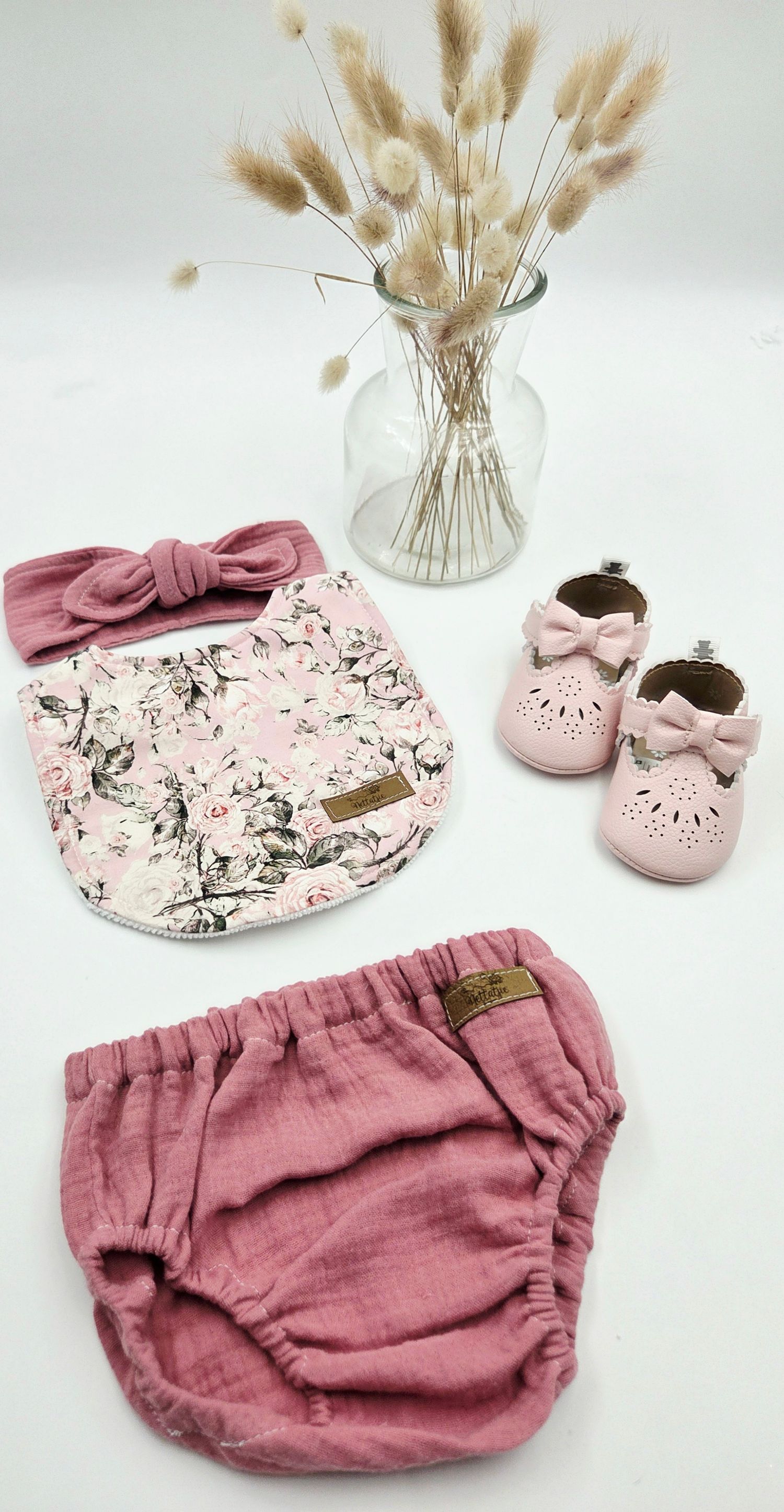 Pink baby bloomers and shoes displayed with dried pampas grass and floral print accessories.