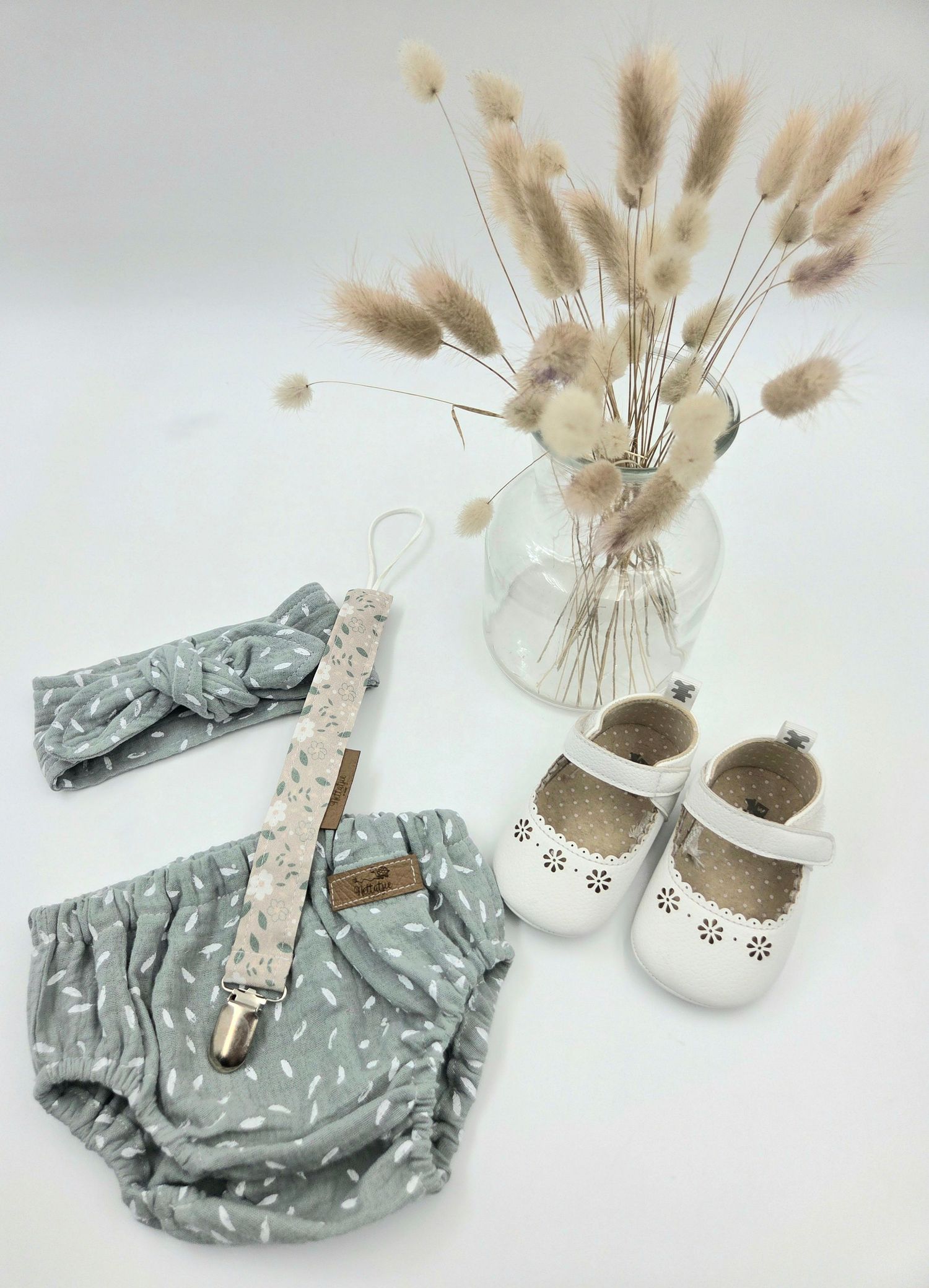 Gray baby bloomers and white shoes arranged with dried pampas grass in a vase.