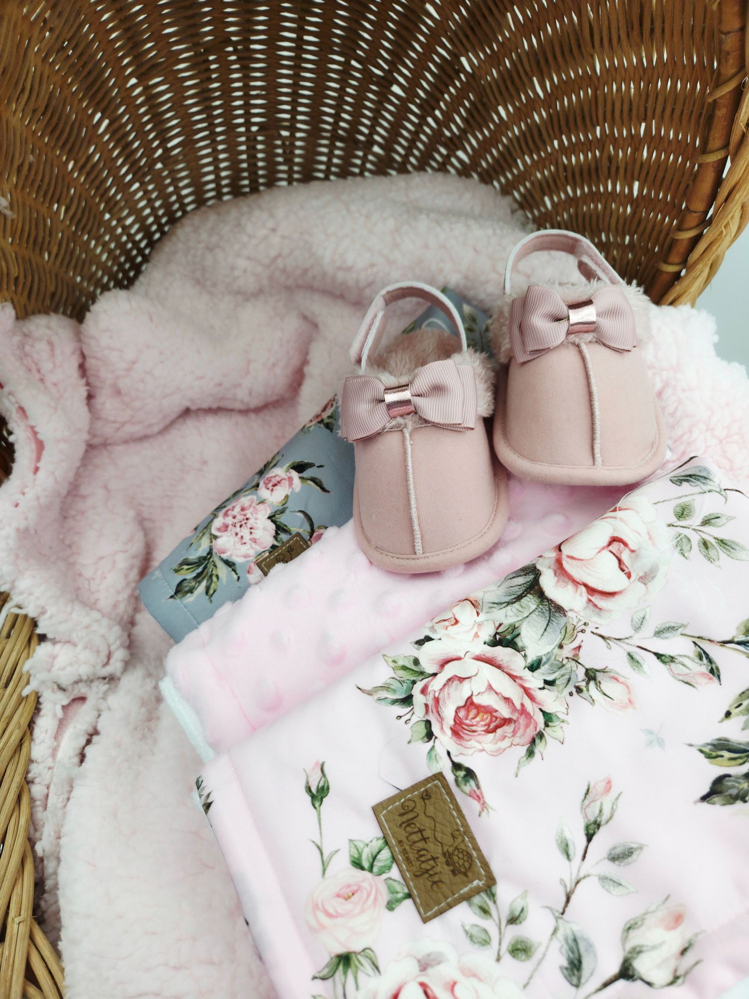 Pink floral baby items and shoes arranged in wicker basket with plush fabric.