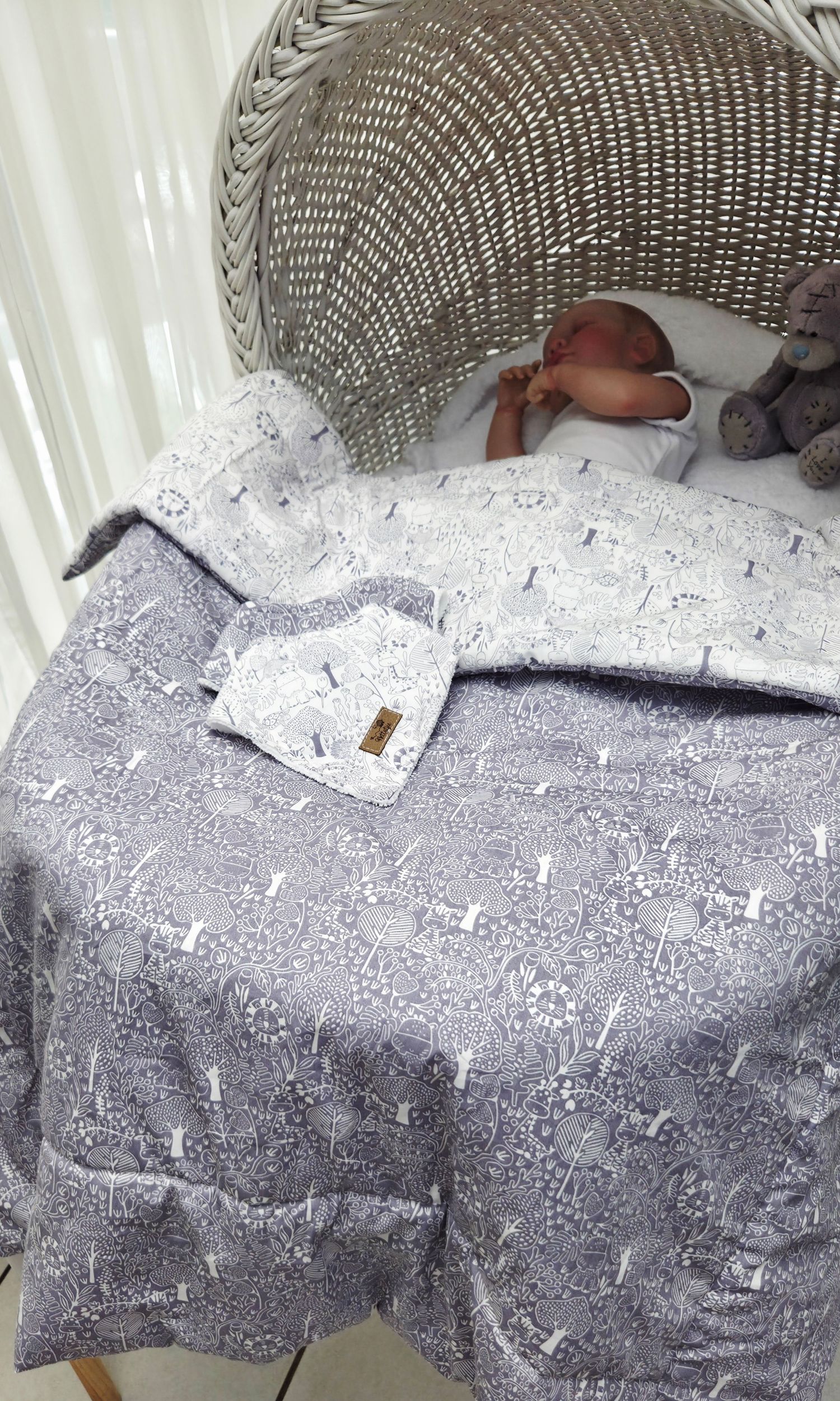 Baby sleeping in a wicker bassinet covered with a gray and white patterned blanket with a matching hat.