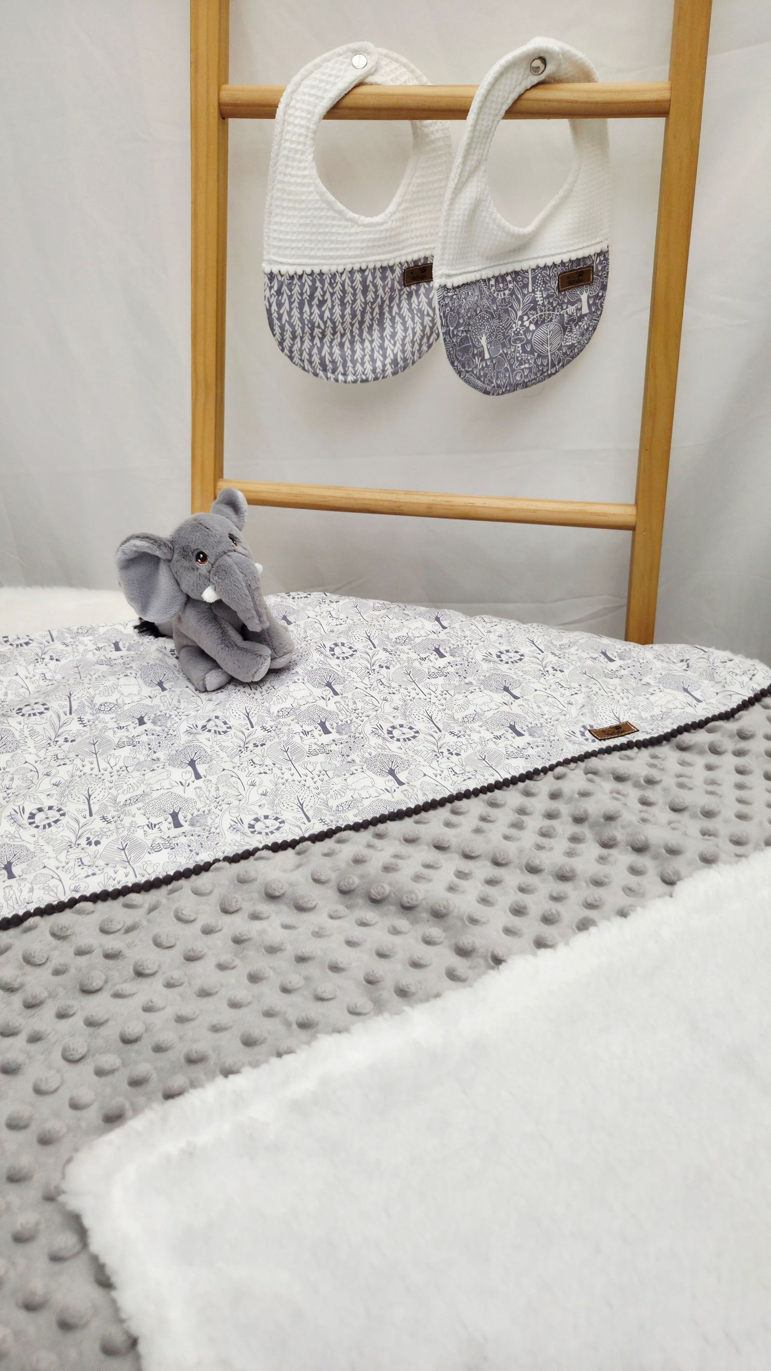 Gray minky baby blanket with elephant plush toy and two white bibs hanging on a wooden ladder display.