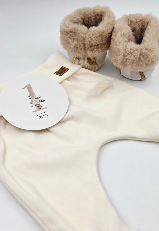 Cream baby bib with 1 week milestone card and fluffy beige fur booties on white background.