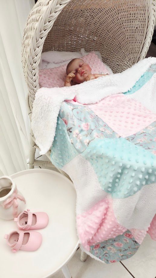 Sleeping newborn baby in white wicker chair covered with pink and teal minky patchwork quilt, pink mary jane shoes on table.