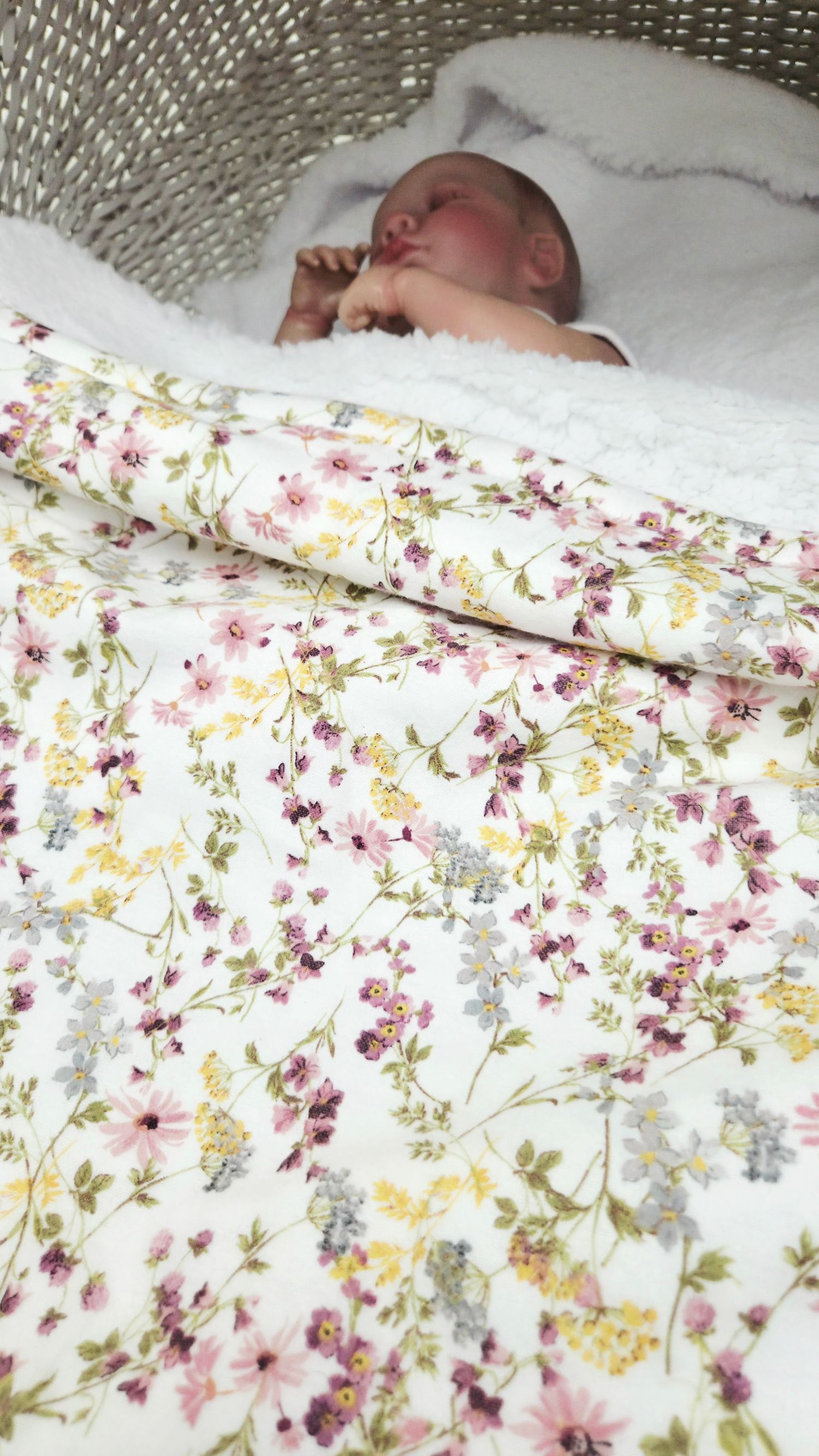 Newborn baby sleeping peacefully in a wicker bassinet with a white floral blanket featuring pink and yellow flowers.
