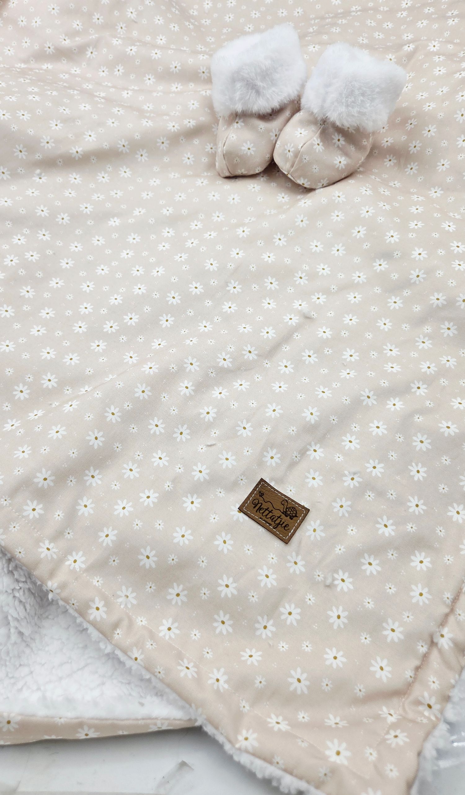 Soft floral baby blanket in blush pink with white daisy pattern, paired with fluffy white baby booties.