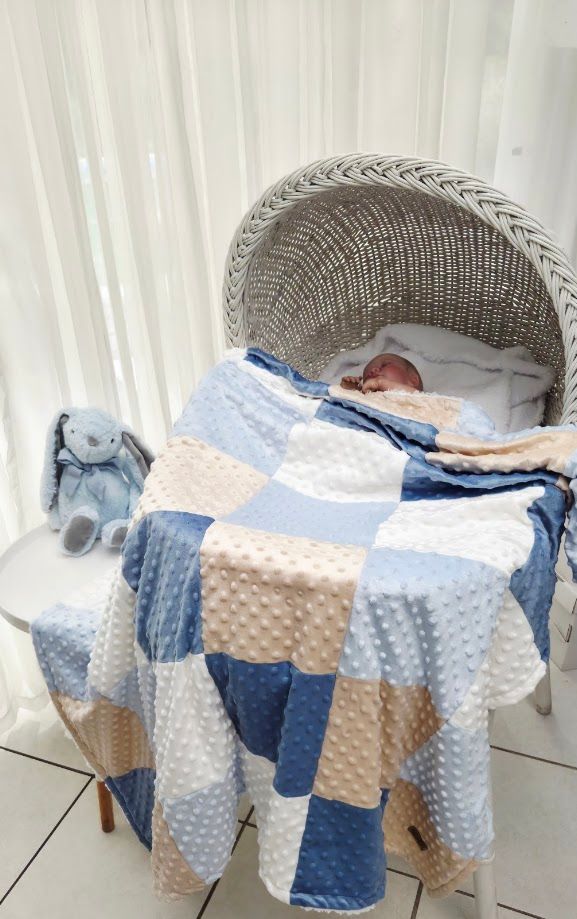 Newborn baby resting in a white wicker bassinet covered with a blue, white, and beige patchwork minky dot blanket.