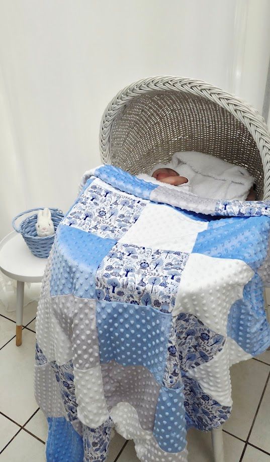 A newborn baby rests in a white wicker bassinet covered with a blue and white patchwork quilt.