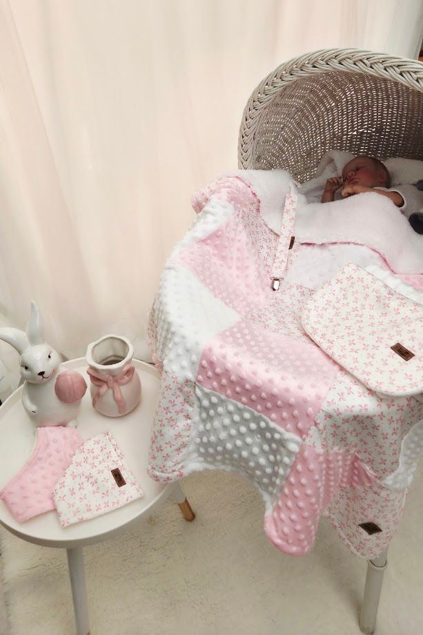 Baby sleeping under a pink patchwork quilt in a wicker bassinet, with booties and bunny decor on a white side table.