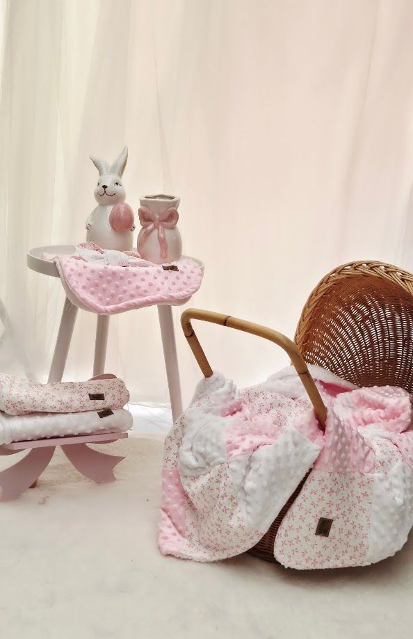 Pink floral baby blanket draped over wicker pram with white bunny figurine and matching bib on small wooden stool.