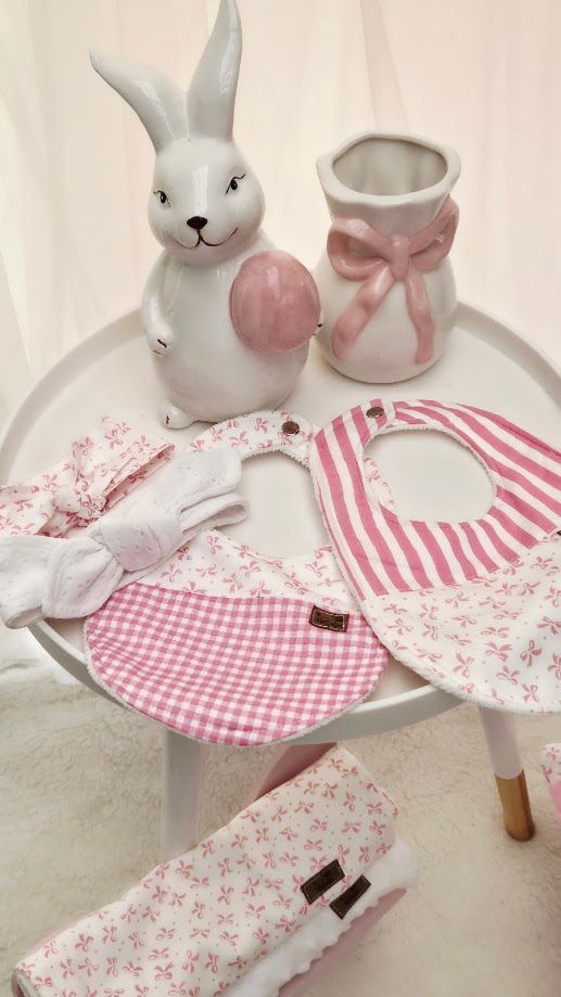 Pink and white baby gift set with bibs, headband, and burp cloth beside ceramic bunny and bow vase décor.