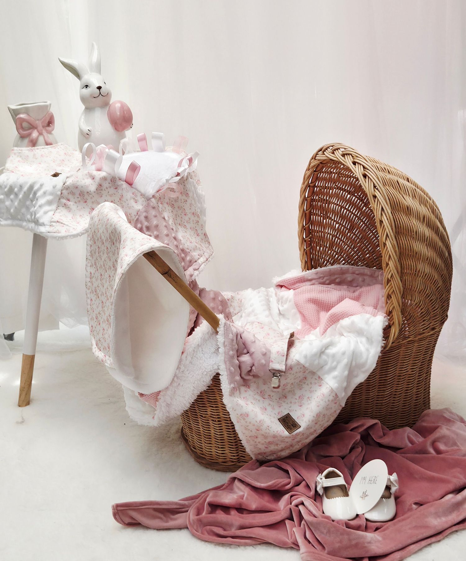 Wicker baby bassinet with pink floral bedding, white bunny toy, baby shoes, and soft blankets in a nursery setting.