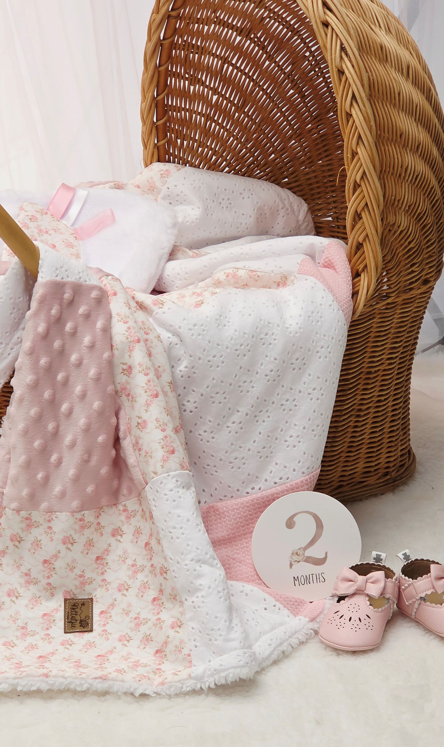 Pink baby quilt with floral and minky fabric draped over wicker bassinet, with 2-month milestone disc and pink bow shoes.