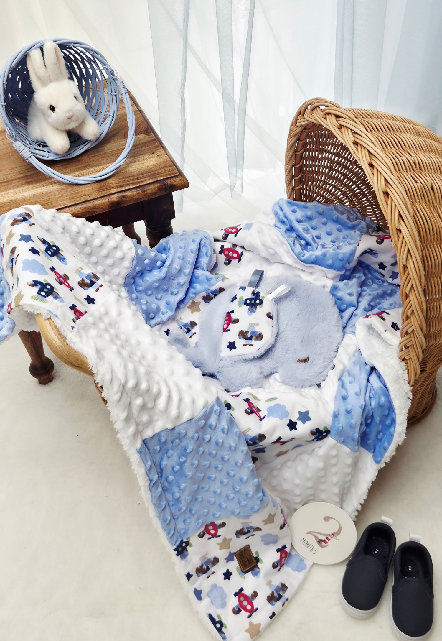 Blue patchwork baby blanket with airplane print, minky fabric, matching bib, stuffed bunny, wicker basket, and black shoes.