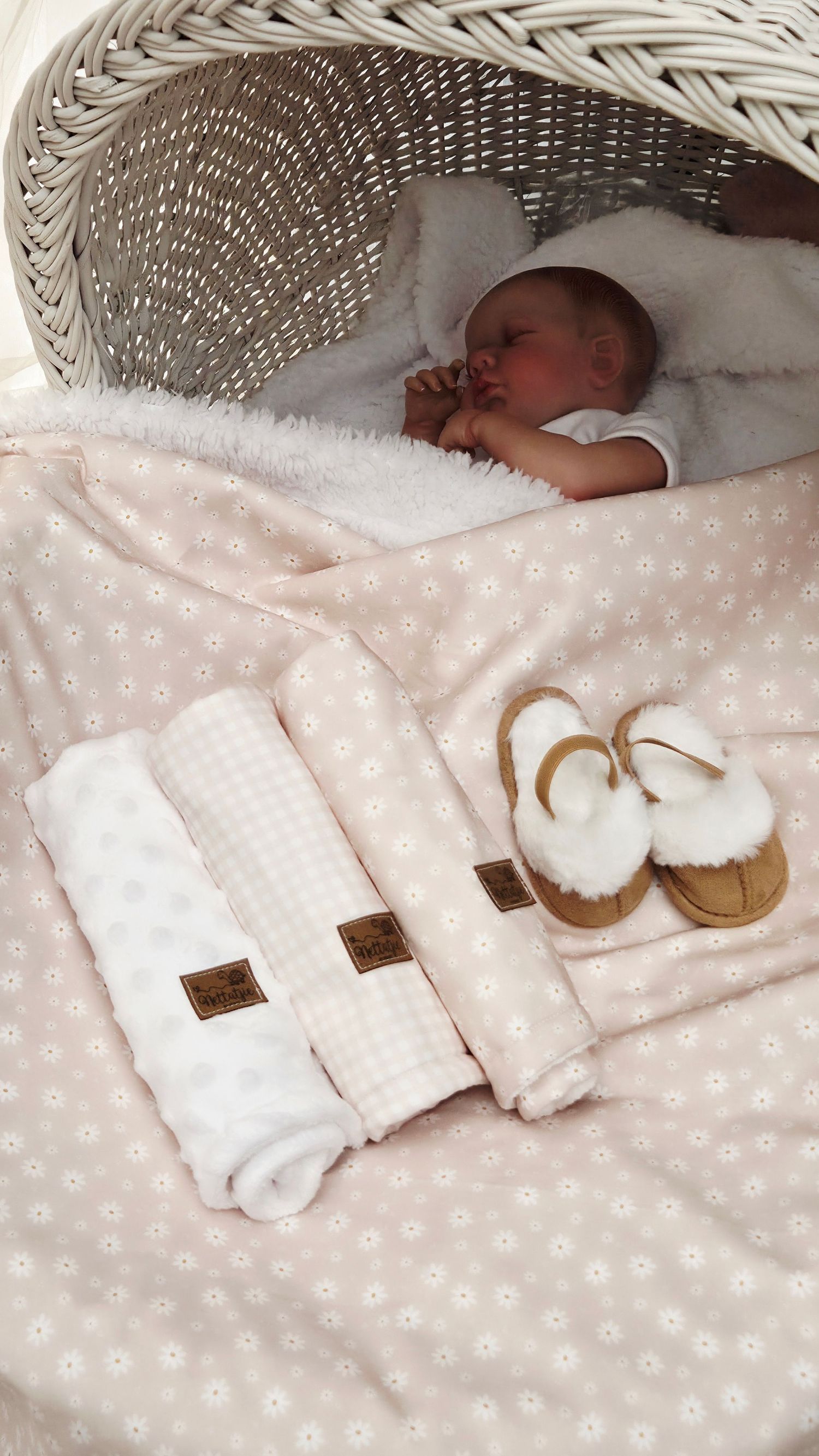 Newborn baby sleeping in wicker bassinet with pink floral blanket, folded muslin cloths, and tan UGG baby slippers.