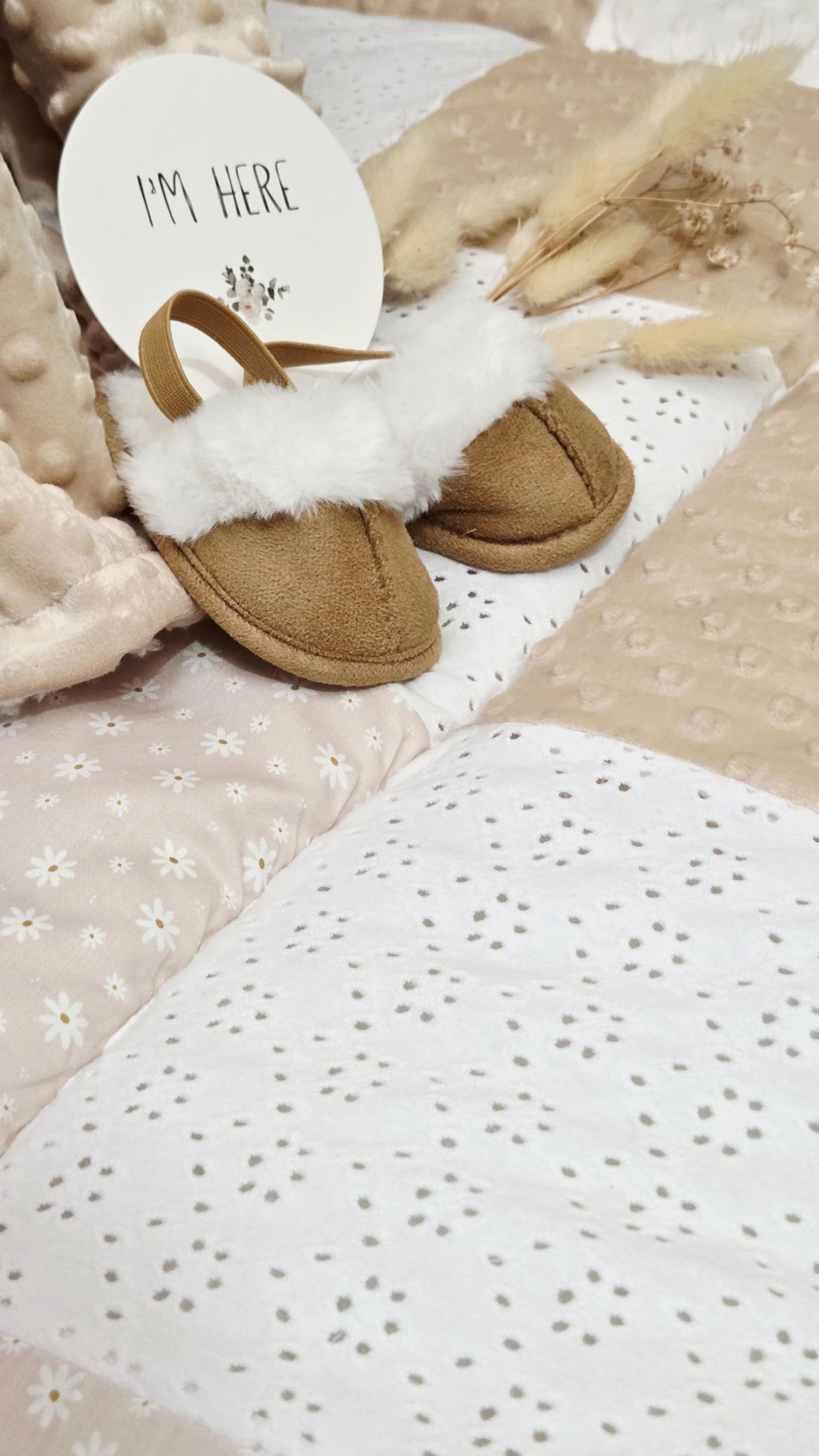 Tiny tan fur-trimmed baby slippers beside an I'm Here sign on daisy print fabric with dried pampas grass.