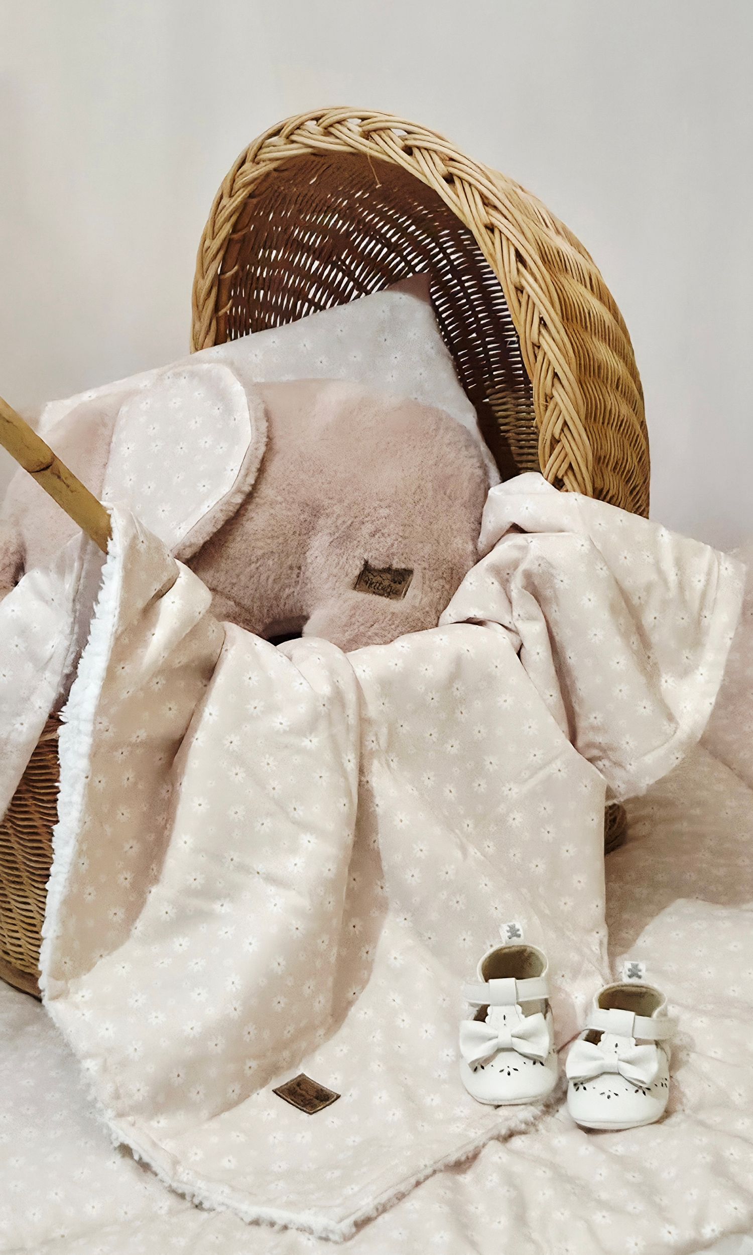 Pink baby blanket with bunny ears and white bow shoes beside a wicker basket, perfect for a newborn gift set.