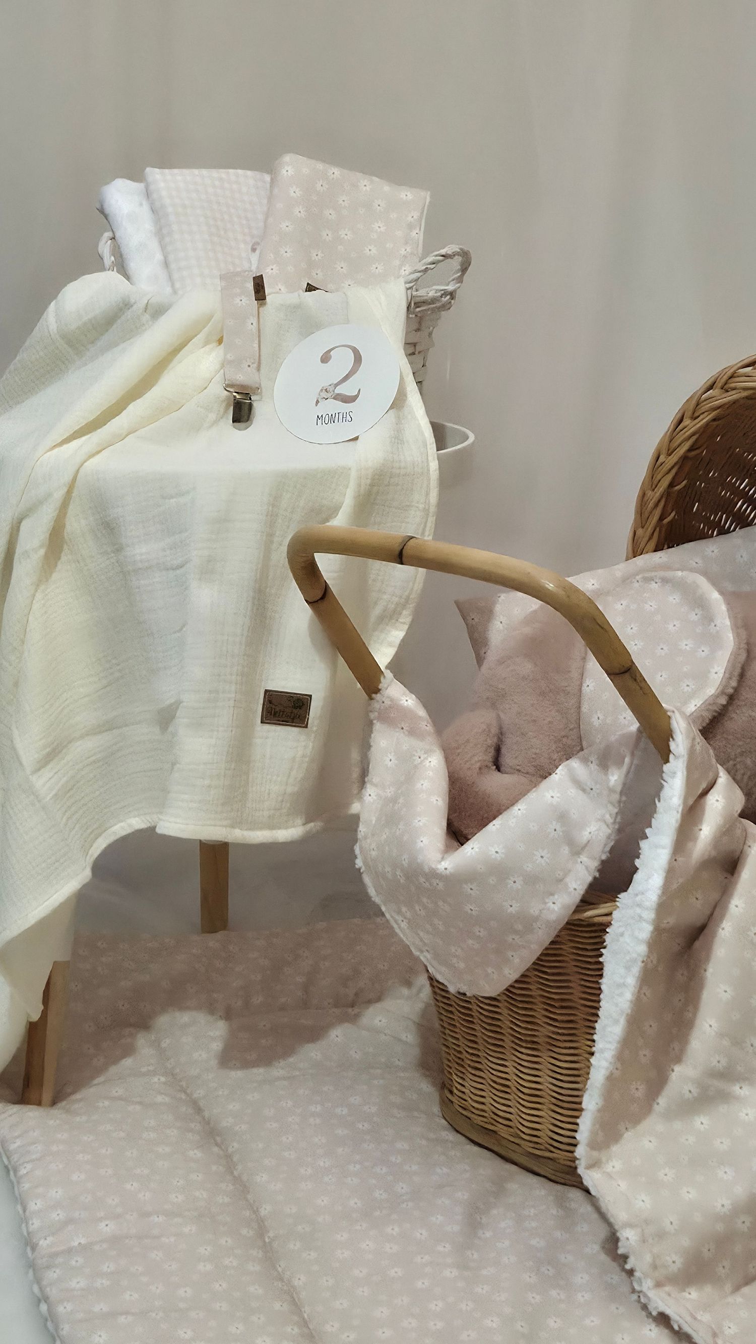Soft cream and blush baby blankets draped over a wooden stool beside a wicker basket with pink sherpa lining.