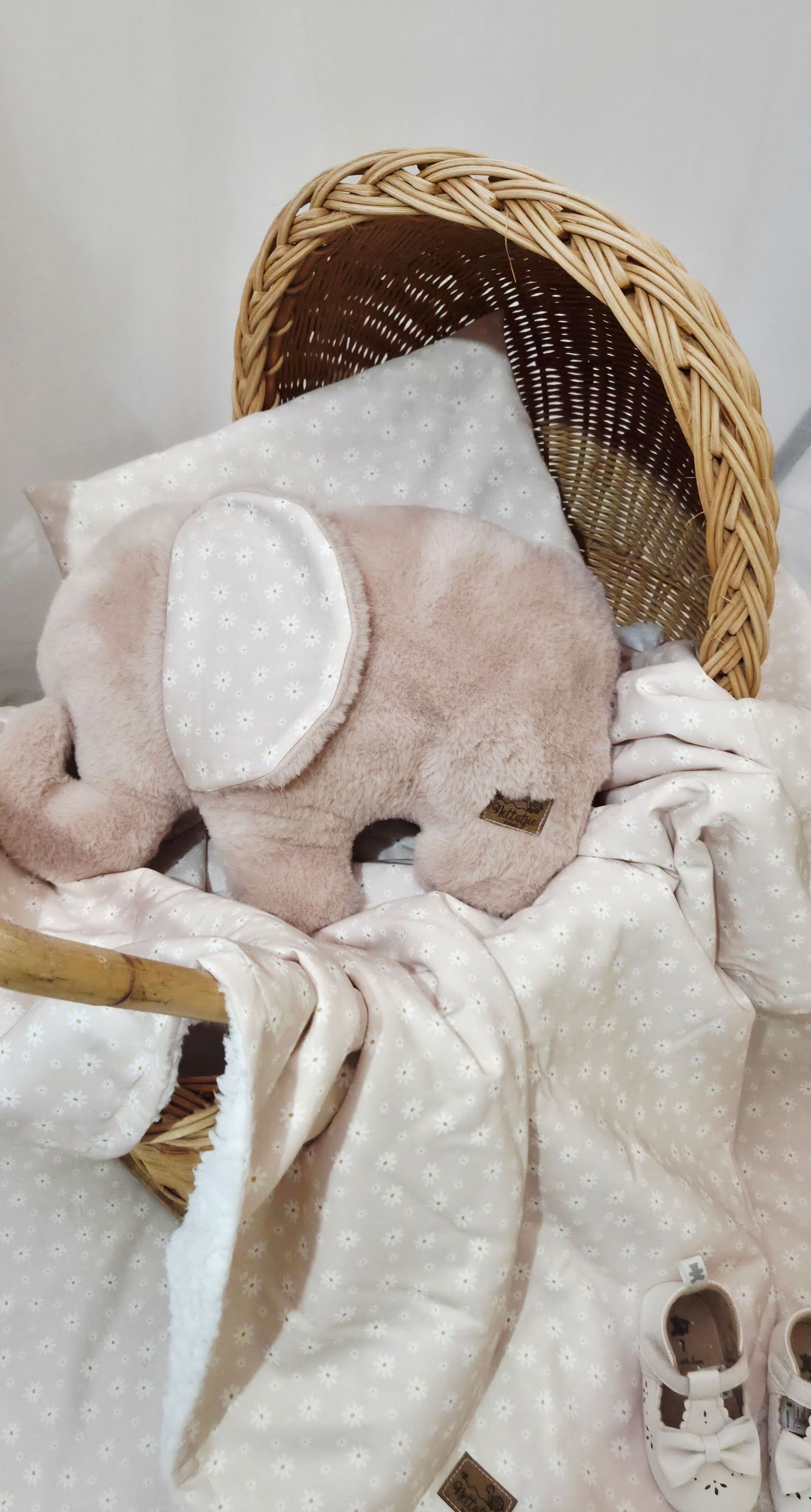 Pink elephant plush toy with floral blanket and baby shoes in a wicker Moses basket.