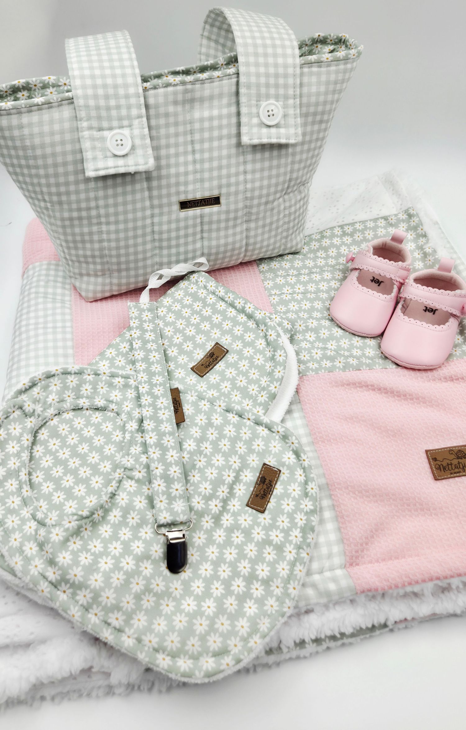 Gray gingham diaper bag set with floral bibs, pink baby shoes, and soft blanket, perfect for newborns.