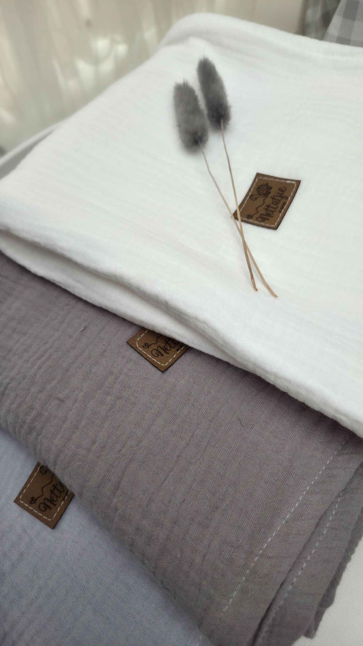Stacked white and gray muslin blankets with leather labels and dried bunny tail grass on top.