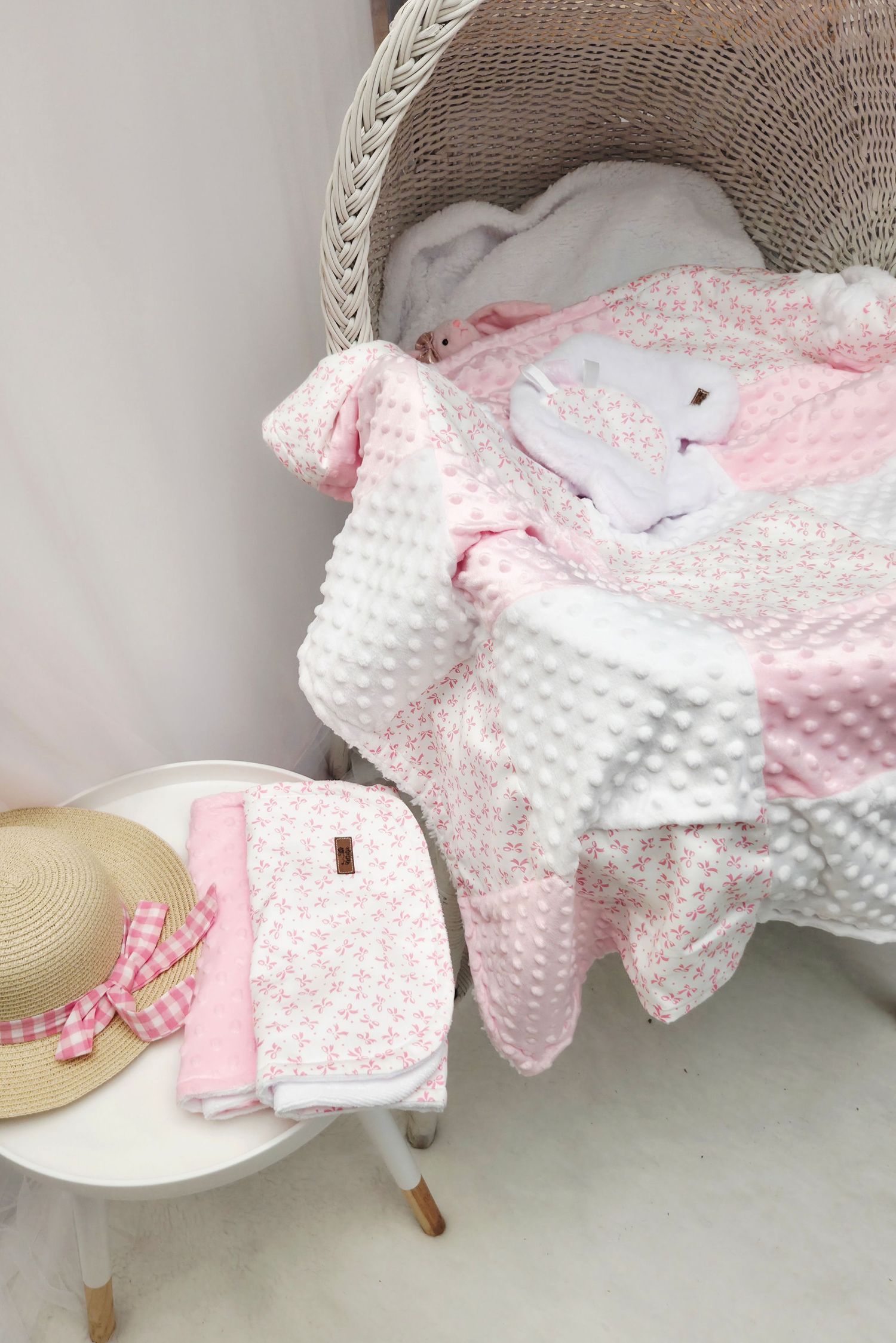 Pink floral baby blanket with bunny toy in wicker bassinet, straw hat with gingham ribbon and folded blanket on white stool.