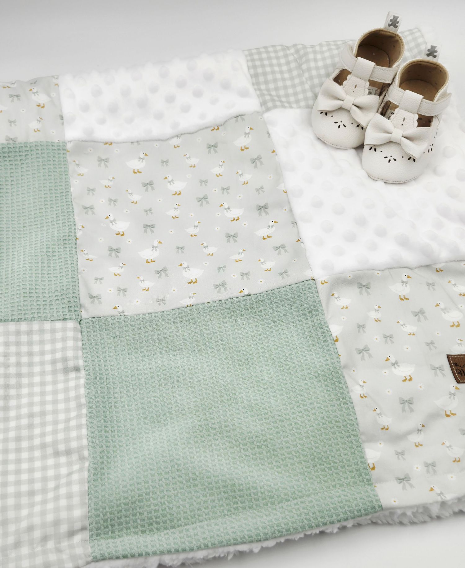 Sage green patchwork baby blanket with lamb print and waffle texture squares, displayed with white baby shoes.