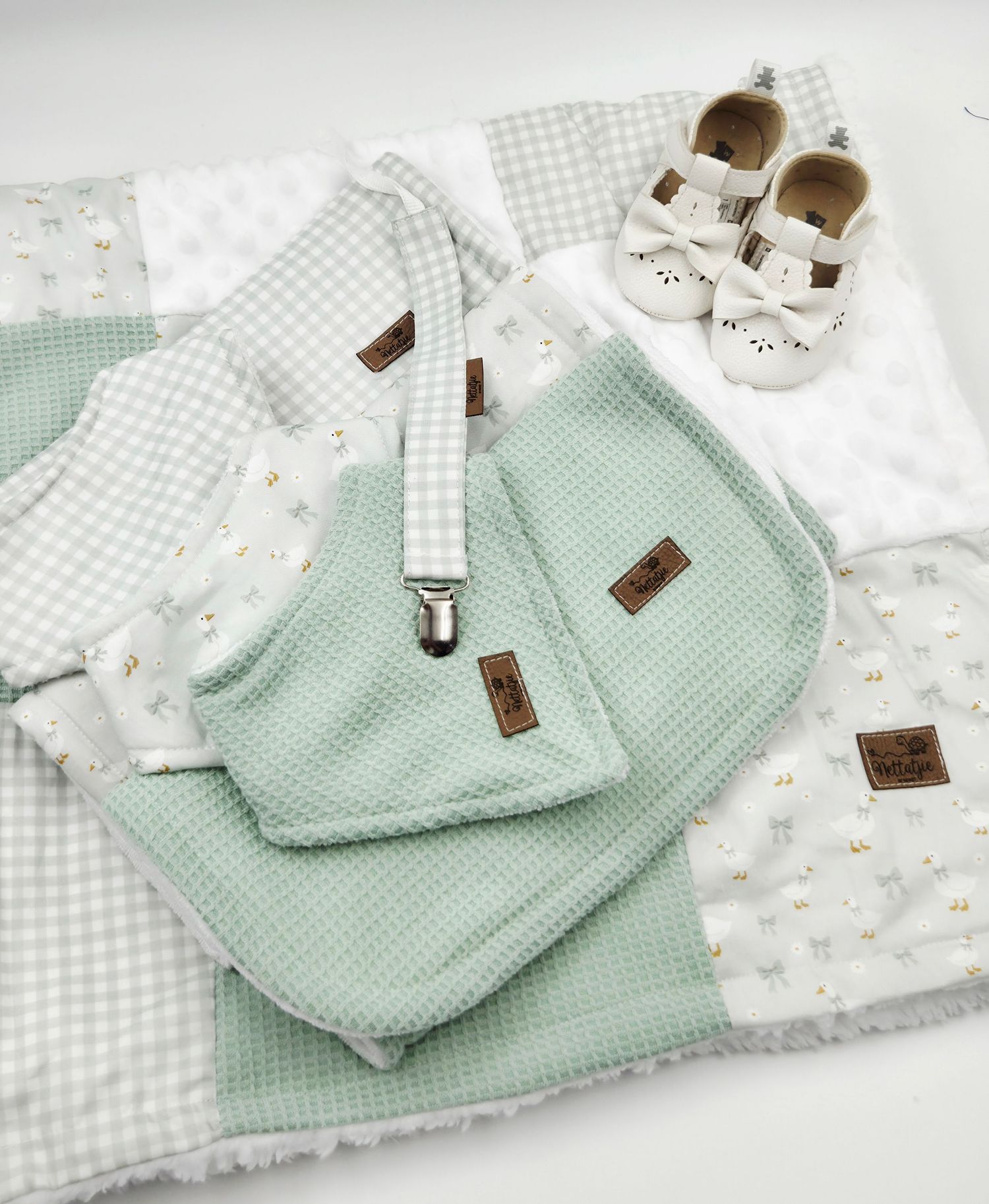 Sage green baby gift set with patchwork blanket, waffle cloth, pacifier clip, and white shoes.