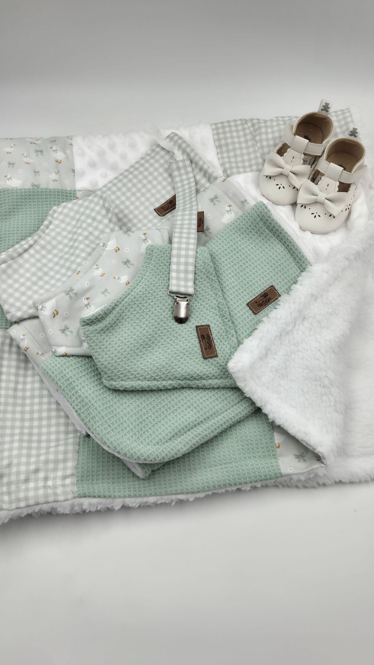 Sage green baby gift set with patchwork blanket, waffle bib, pacifier clip, and white shoes.