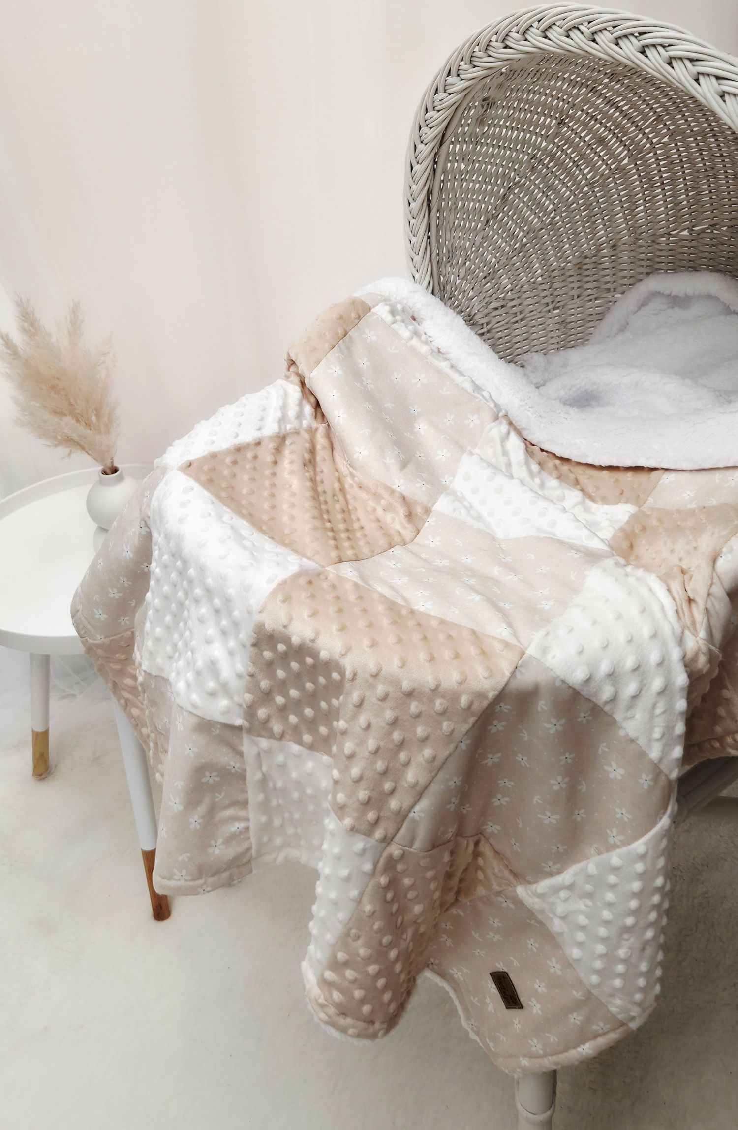 Neutral beige and white patchwork minky baby blanket draped over a white wicker chair with pampas grass decor.