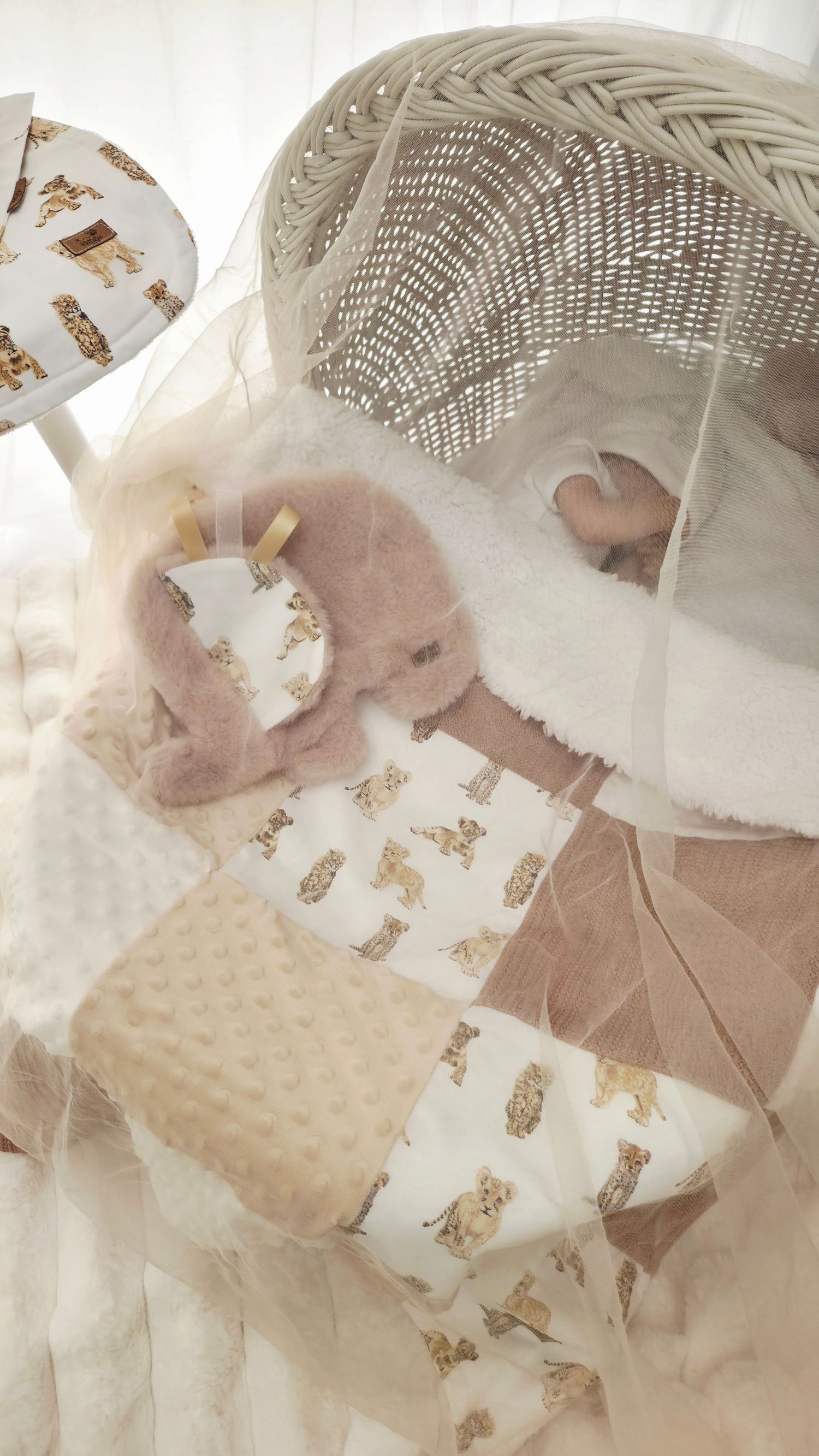 Newborn baby sleeping in a wicker bassinet with a teddy bear print quilt and soft plush toy under a tulle canopy.