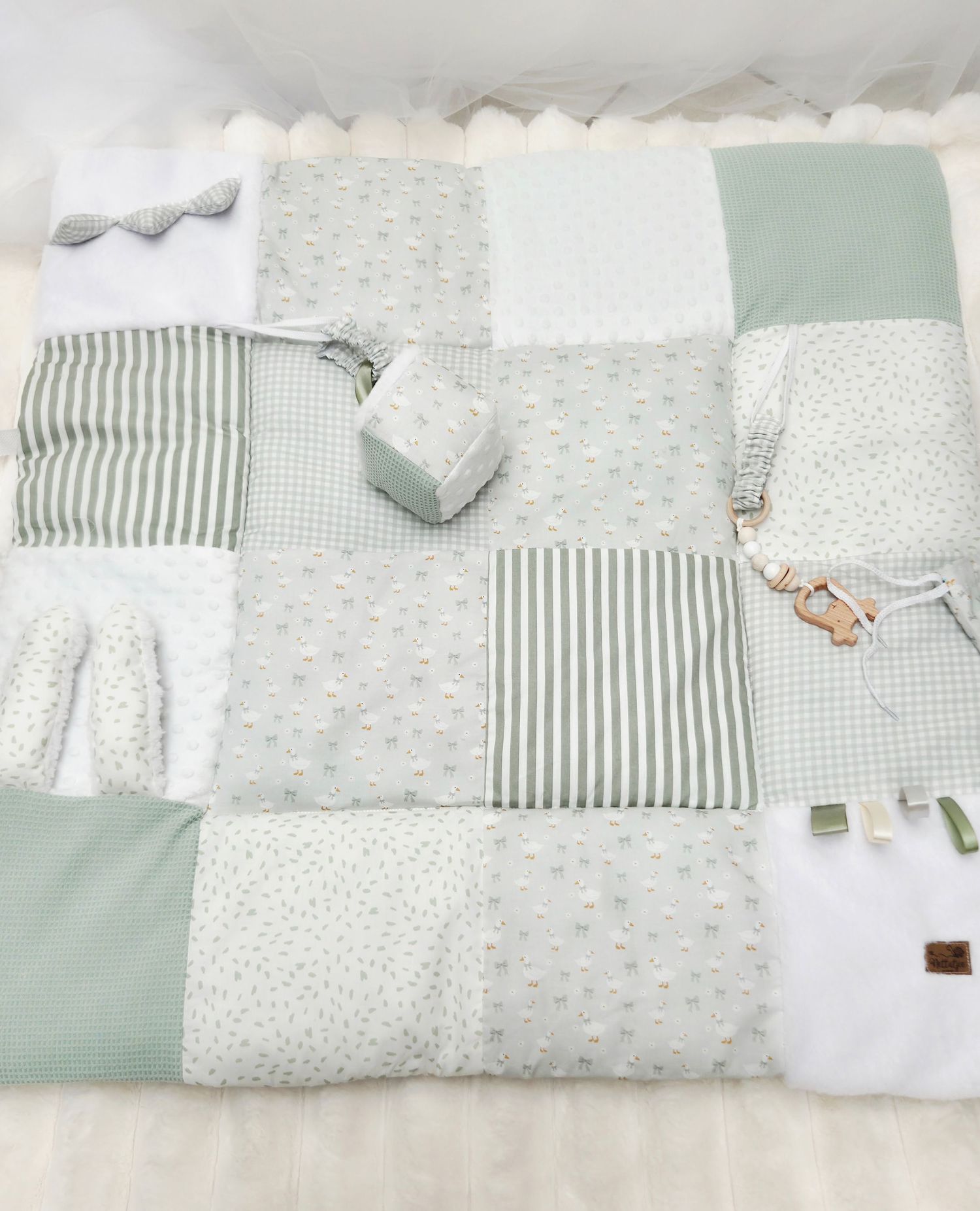Sage green and white patchwork baby play mat with stripes, dots, and gingham patterns, featuring small toys.
