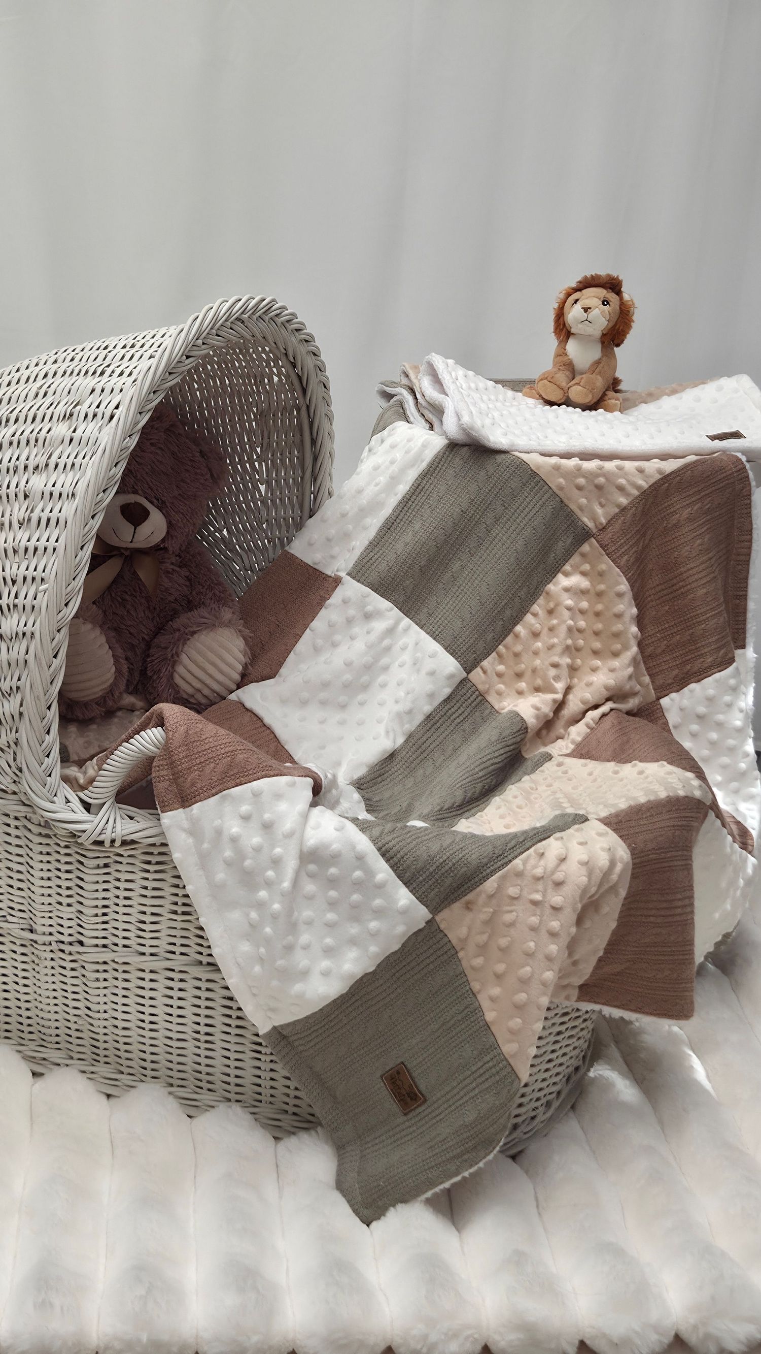 Neutral-toned patchwork baby quilt in white, beige, and sage green with a lion stuffed animal beside a wicker bassinet.