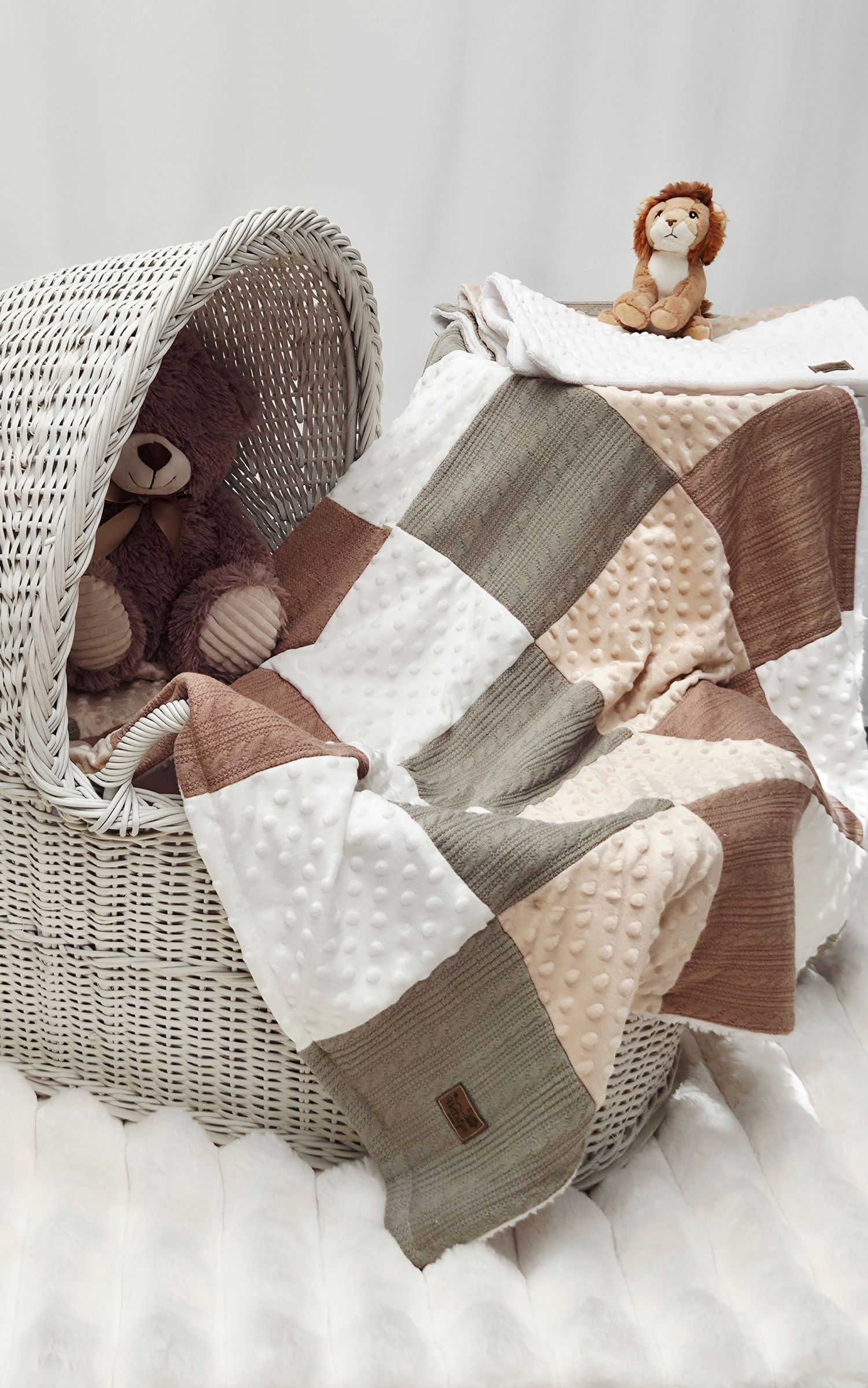 Neutral-toned patchwork baby quilt draped over a wicker bassinet with plush teddy bear and lion stuffed animals.
