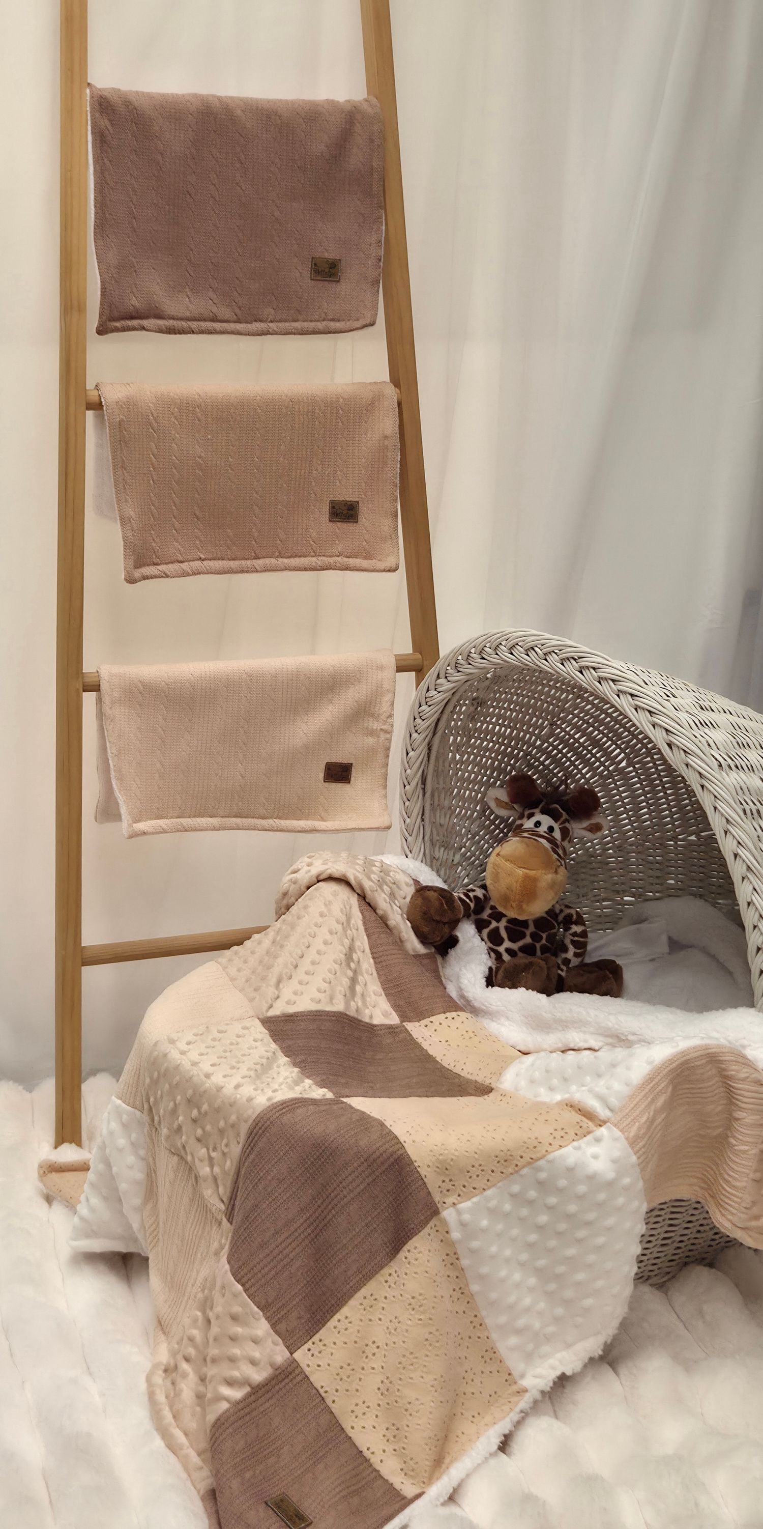 Neutral-toned baby blankets on wooden ladder with patchwork quilt and giraffe plush toy beside wicker bassinet.