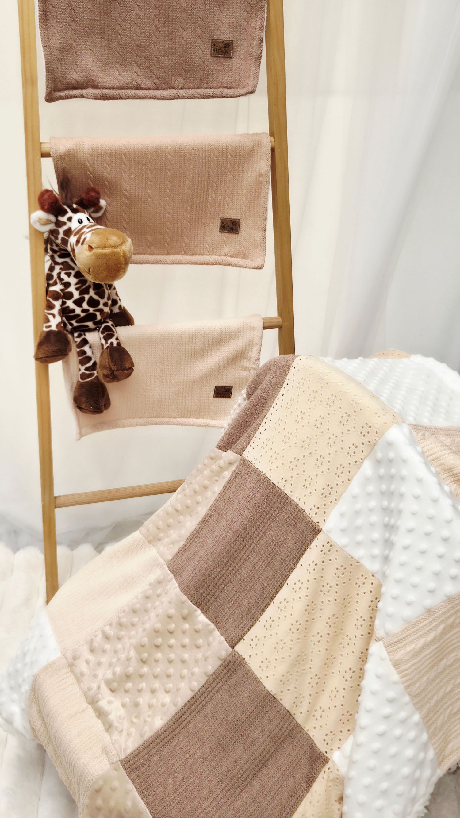 Neutral beige patchwork baby quilt with minky dots beside a wooden ladder holding soft blankets and a giraffe plush toy.
