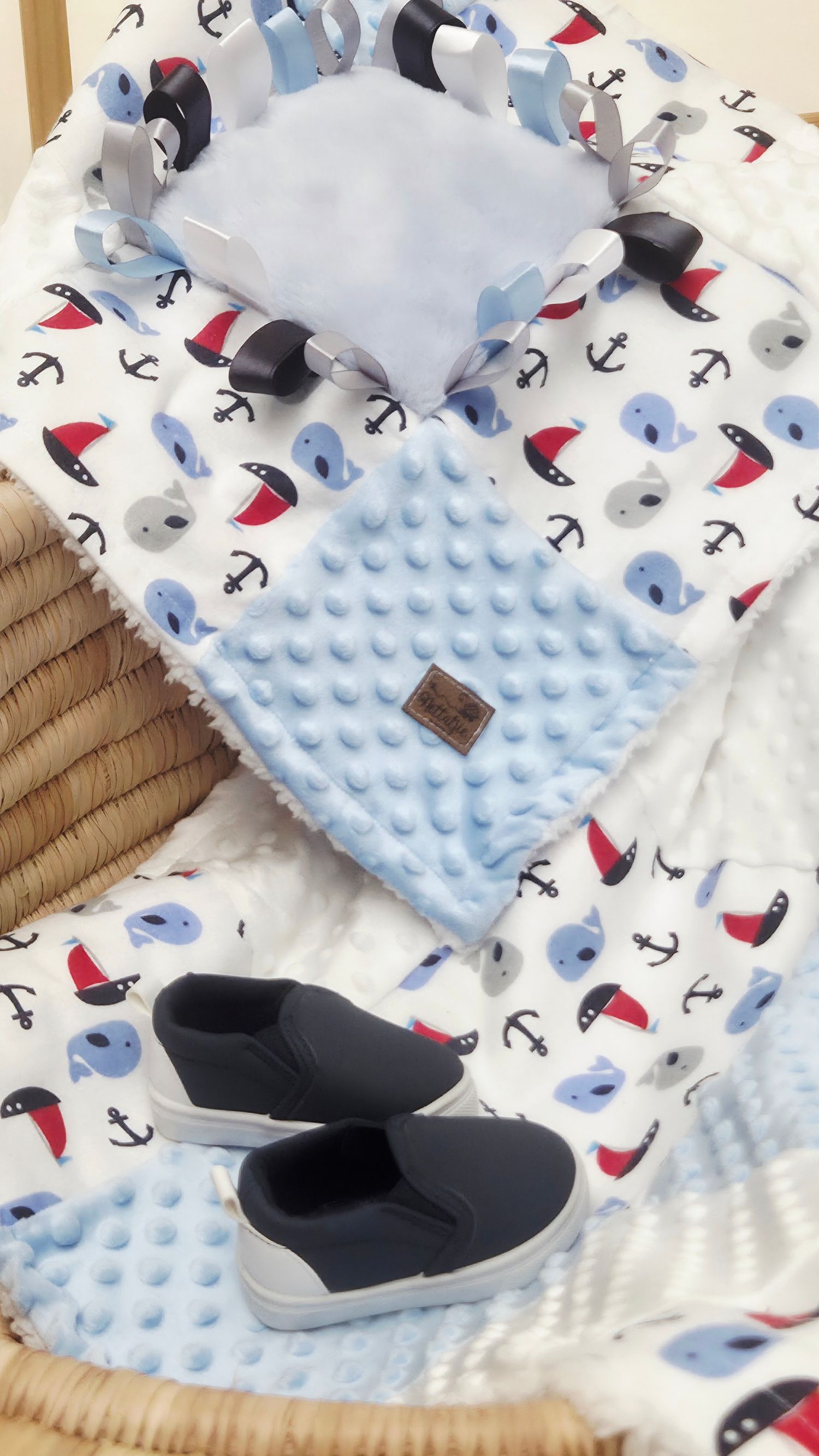 Nautical baby blanket set with sailboat anchor print, blue minky dot lovey, and navy slip-on shoes in a wicker basket.