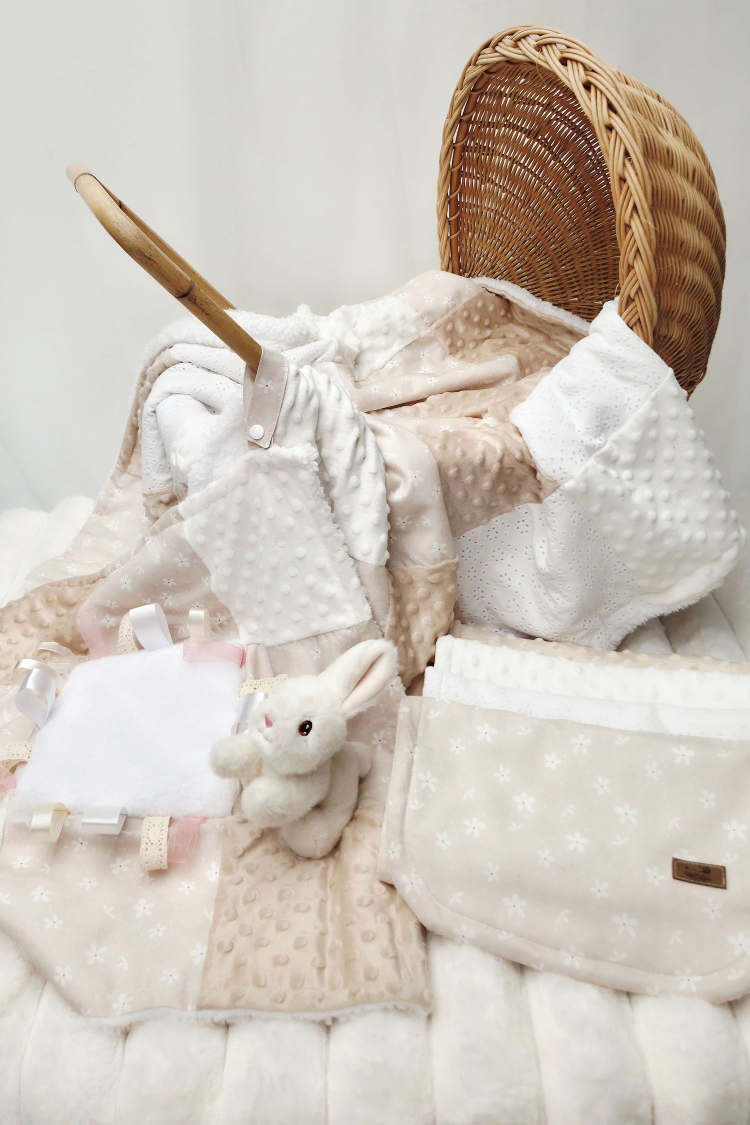Neutral-toned baby gift set with wicker basket, soft blankets, plush bunny toy, and white clothing items.