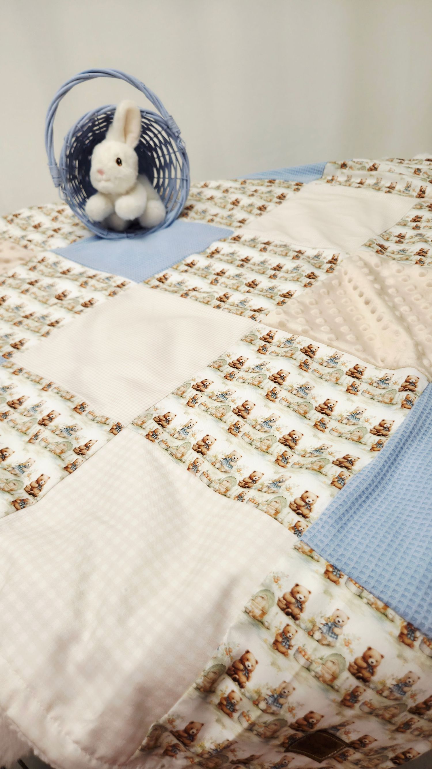Blue and white patchwork baby quilt with teddy bear print, displayed with a white plush bunny in a blue wicker basket.