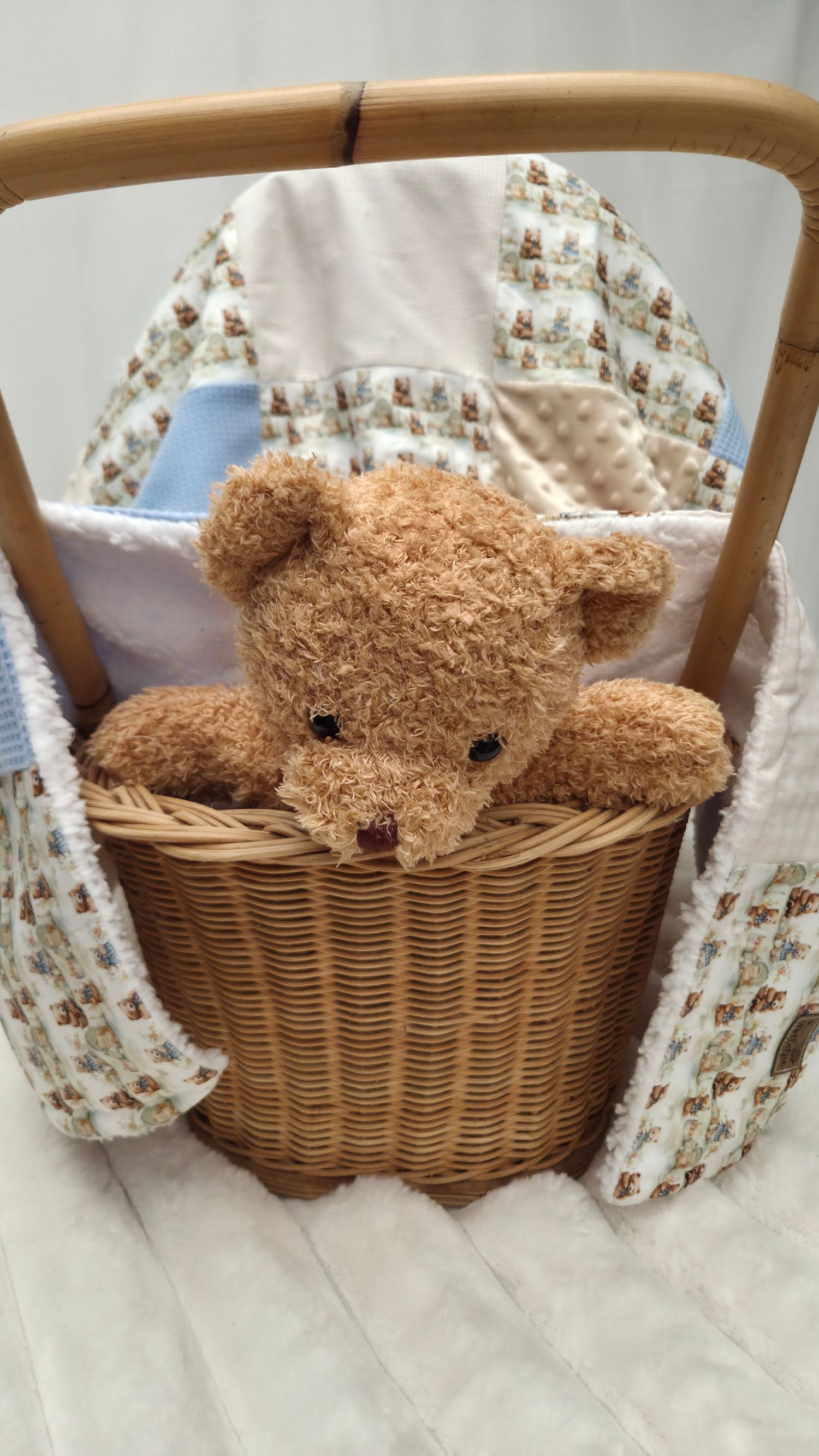 A fluffy brown teddy bear sits in a wicker basket surrounded by soft baby blankets with bear print fabric.