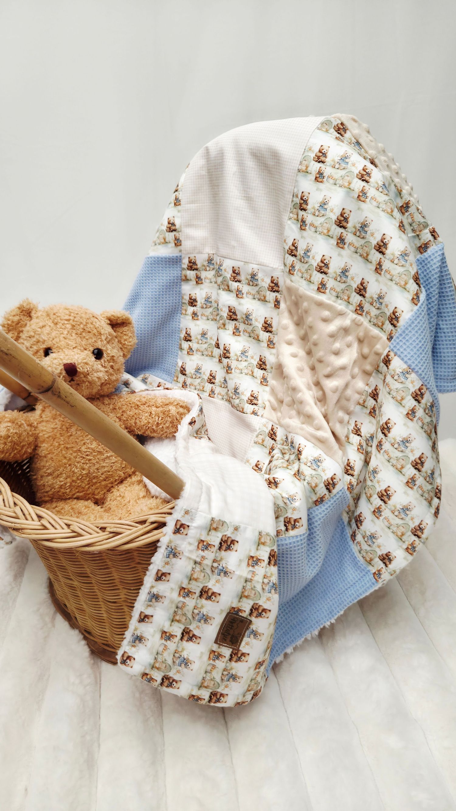 Baby quilt with teddy bear print and blue gingham patches draped over a wicker basket with a plush teddy bear.