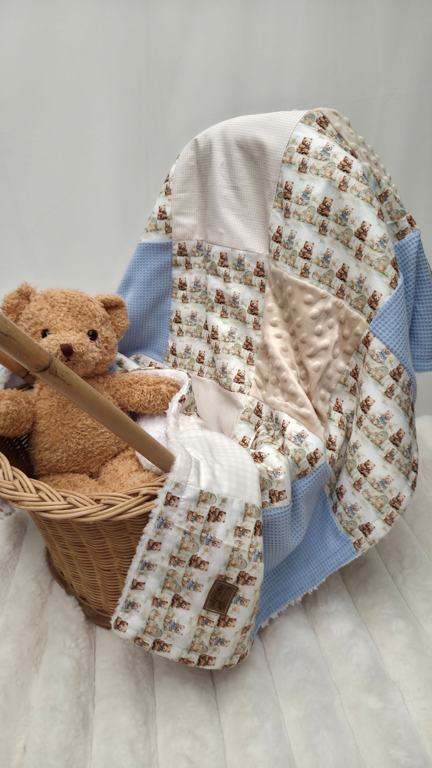 Handmade baby patchwork quilt in blue and white with teddy bear print, displayed with a plush bear in a wicker basket.