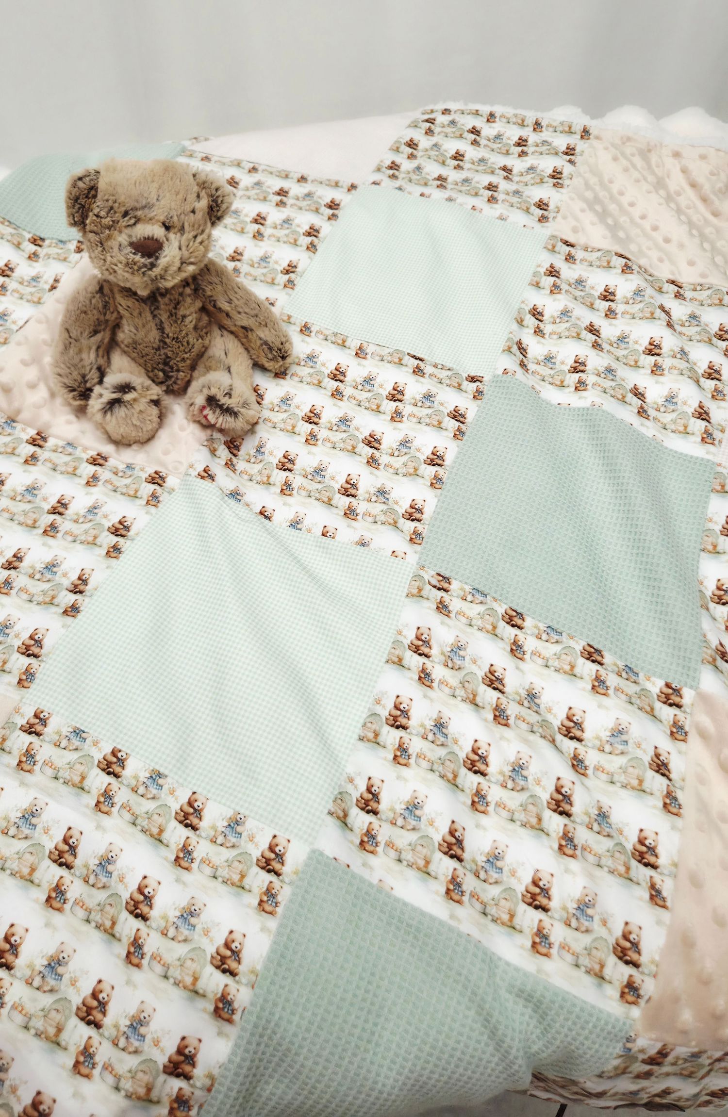 A handmade patchwork baby quilt with mint green and teddy bear print squares, displayed with a small brown stuffed bear toy.