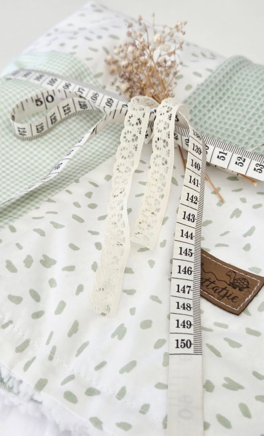 Sewing supplies flat lay with measuring tape, lace trim, green gingham fabric, and spotted white cloth on white background.
