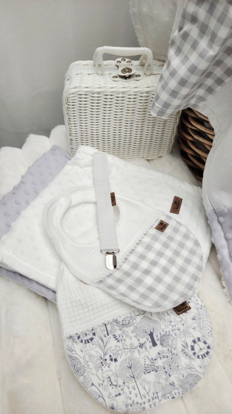 White wicker suitcase with gray gingham baby bib, burp cloth, and floral fabric items arranged on a white surface.